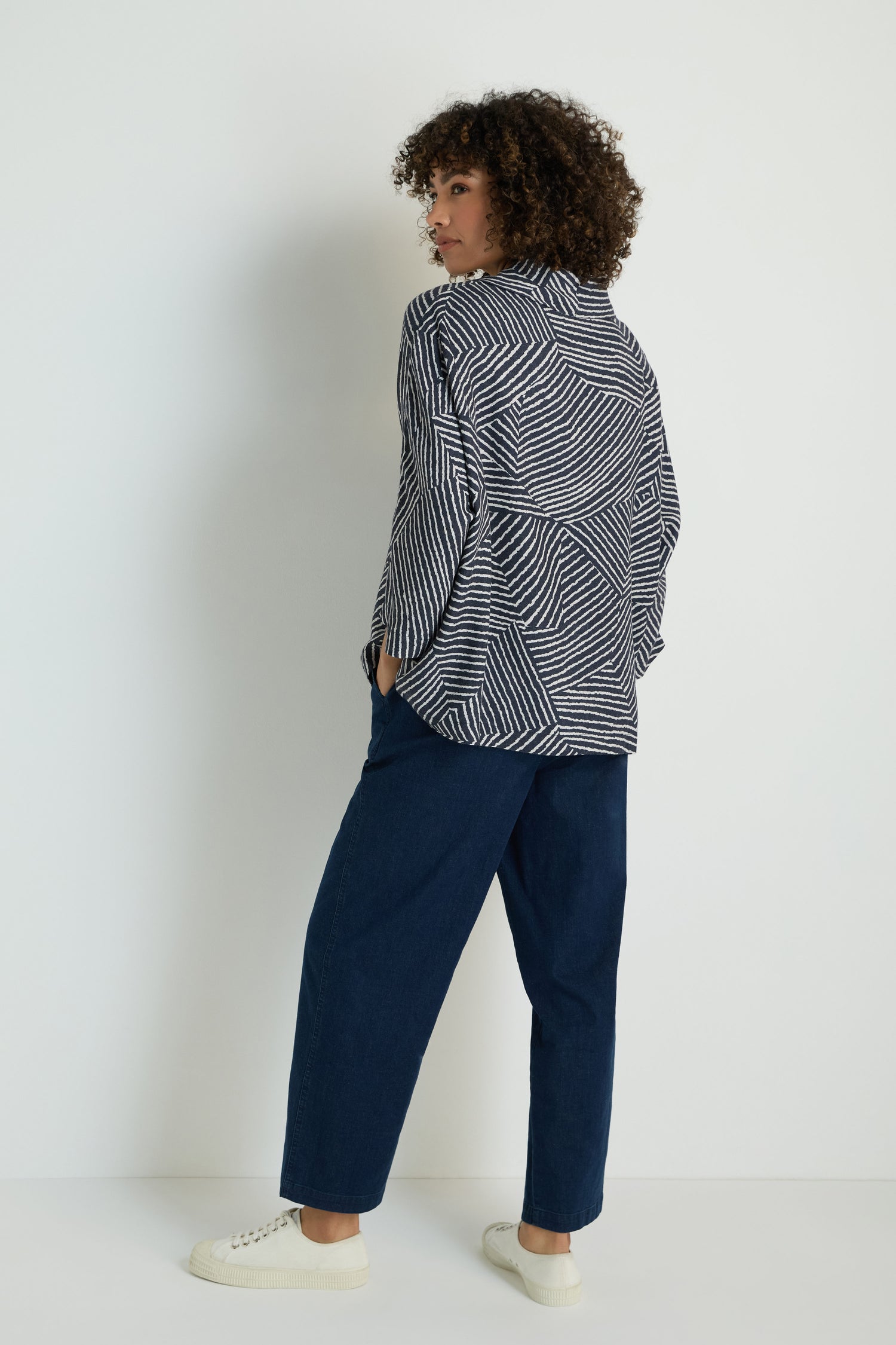 A woman stands facing away, wearing the Linear Prism Lines Cowl Neck Top in navy and white geometric print, paired with light gray wide-leg pants and white slip-on shoes, all set against a plain white background.