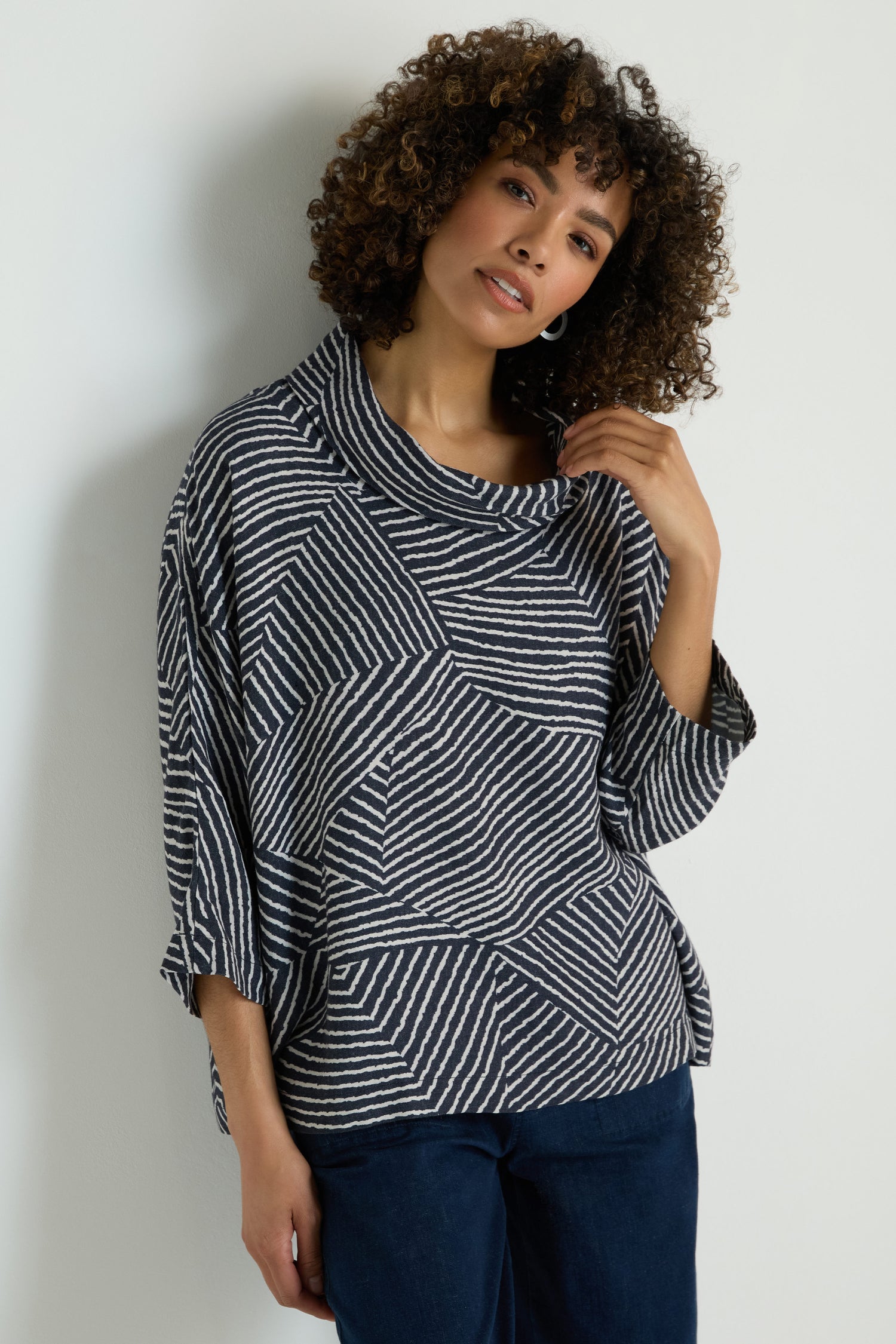 Woman with wavy blond hair wears the Linear Prism Lines Cowl Neck Top, a loose-fitting navy and white geometric print piece crafted from soft brushed viscose, standing against a plain light background.