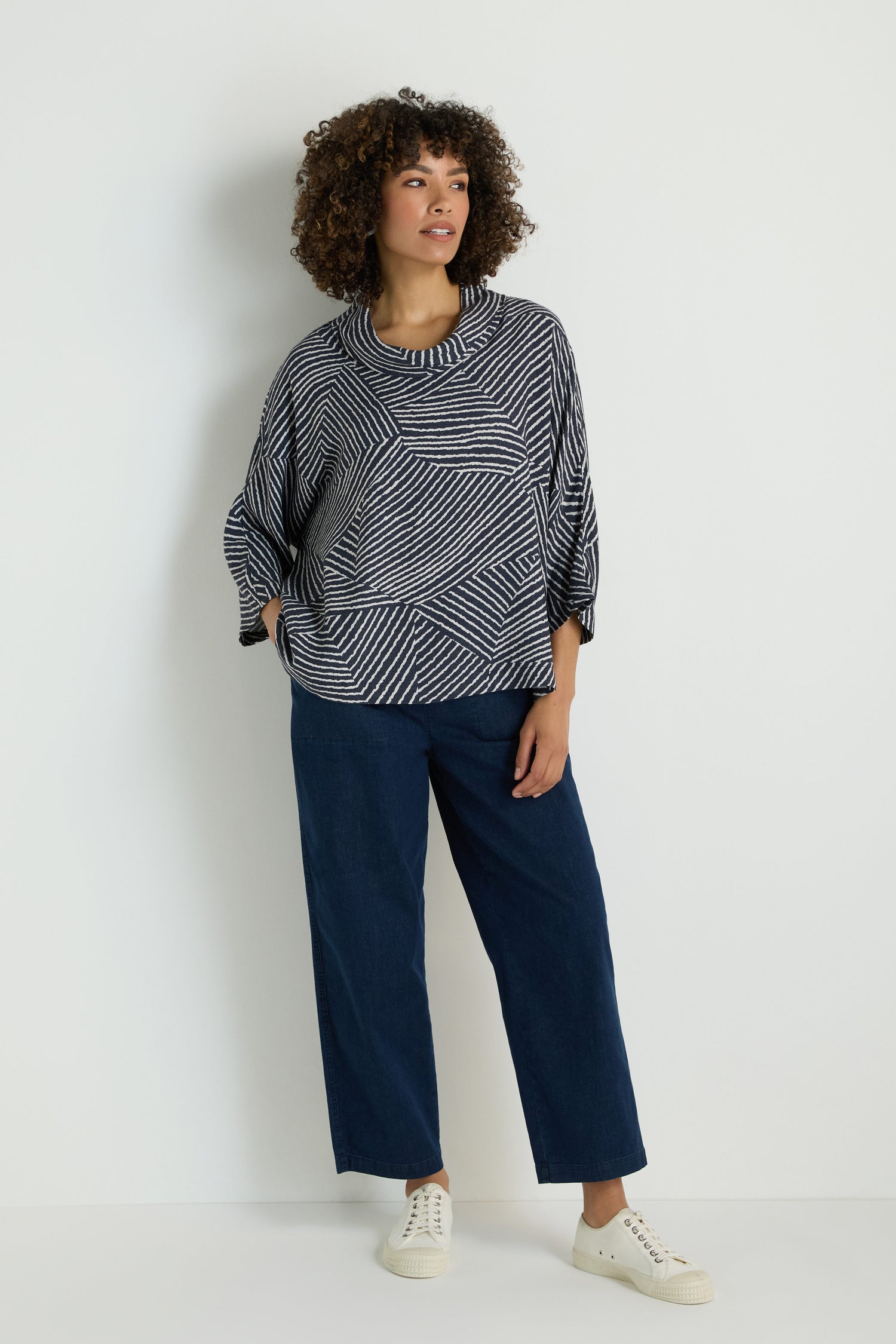 A woman with wavy blonde hair sits on the floor against a plain background, wearing the Linear Prism Lines Cowl Neck Top in navy and white brushed viscose, paired with light gray wide-leg pants and white slip-on sneakers.