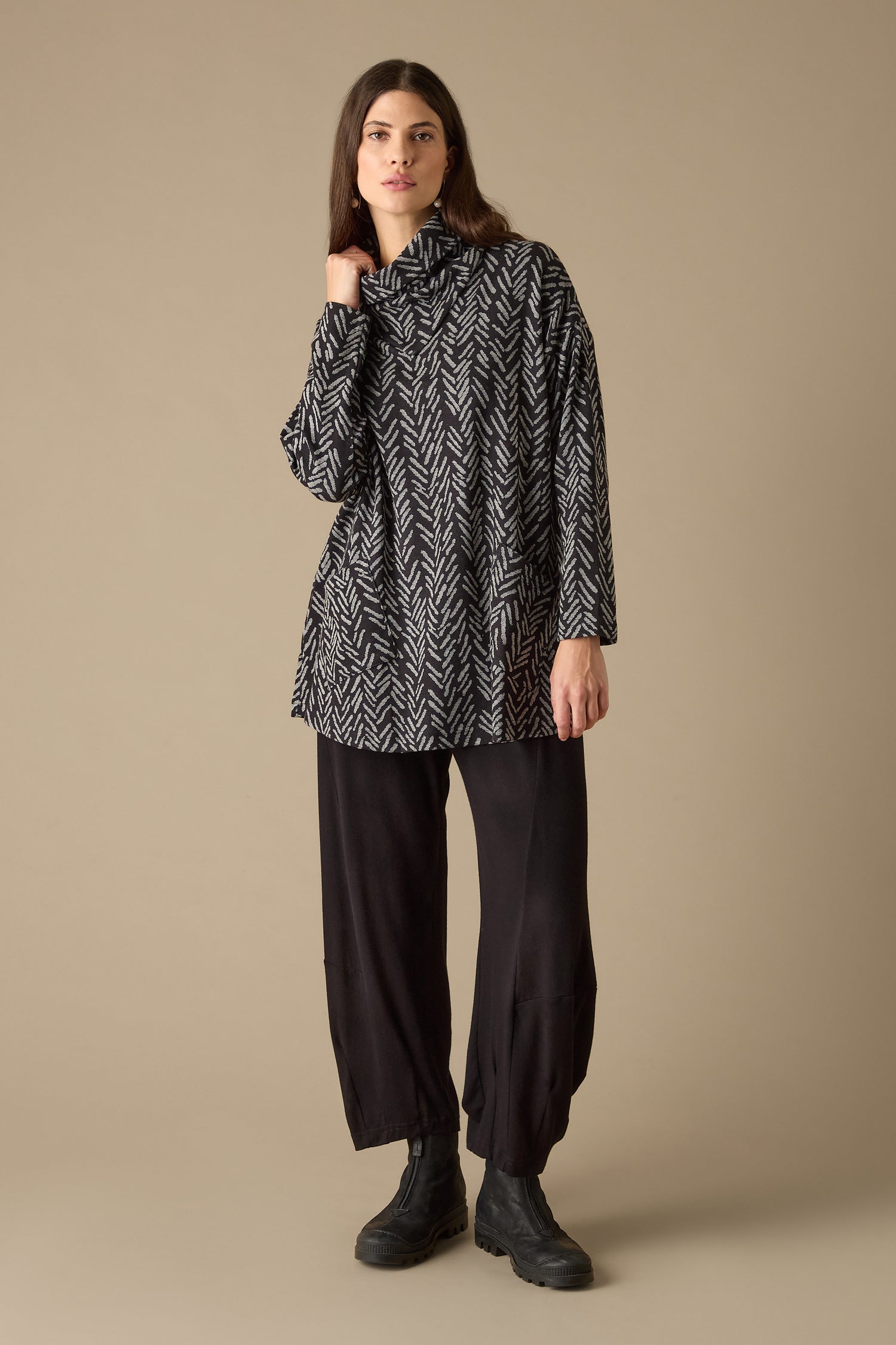 A person dressed in the Herringbone Jersey Jacquard Top with a boxy fit and black pants stands on a beige background, casually holding the funnel neck collar with one hand.