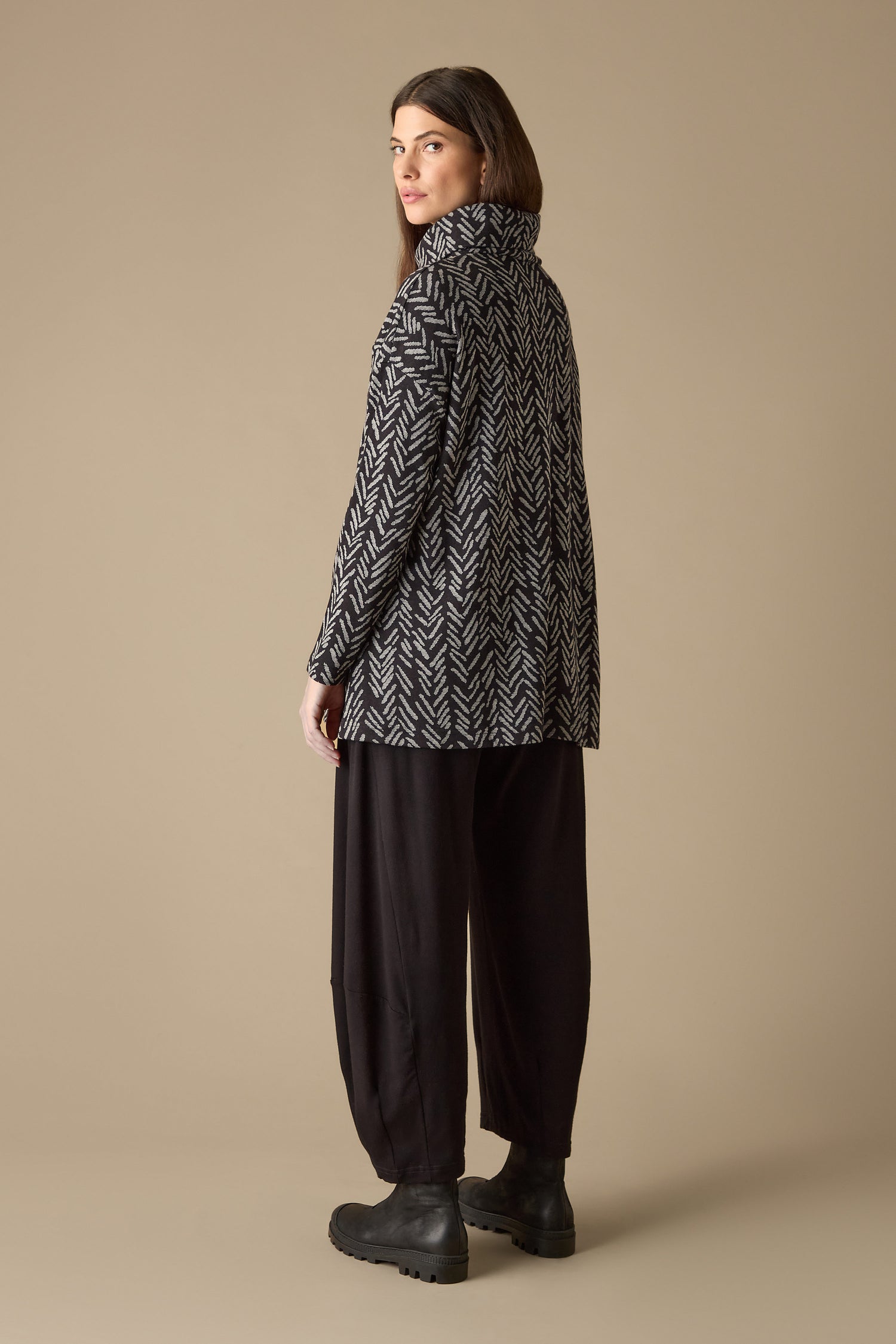 A person dressed in a boxy fit jacket and black pants stands against a neutral background, looking over their shoulder. The outfit is enhanced by the elegant touch of the Herringbone Jersey Jacquard Top.
