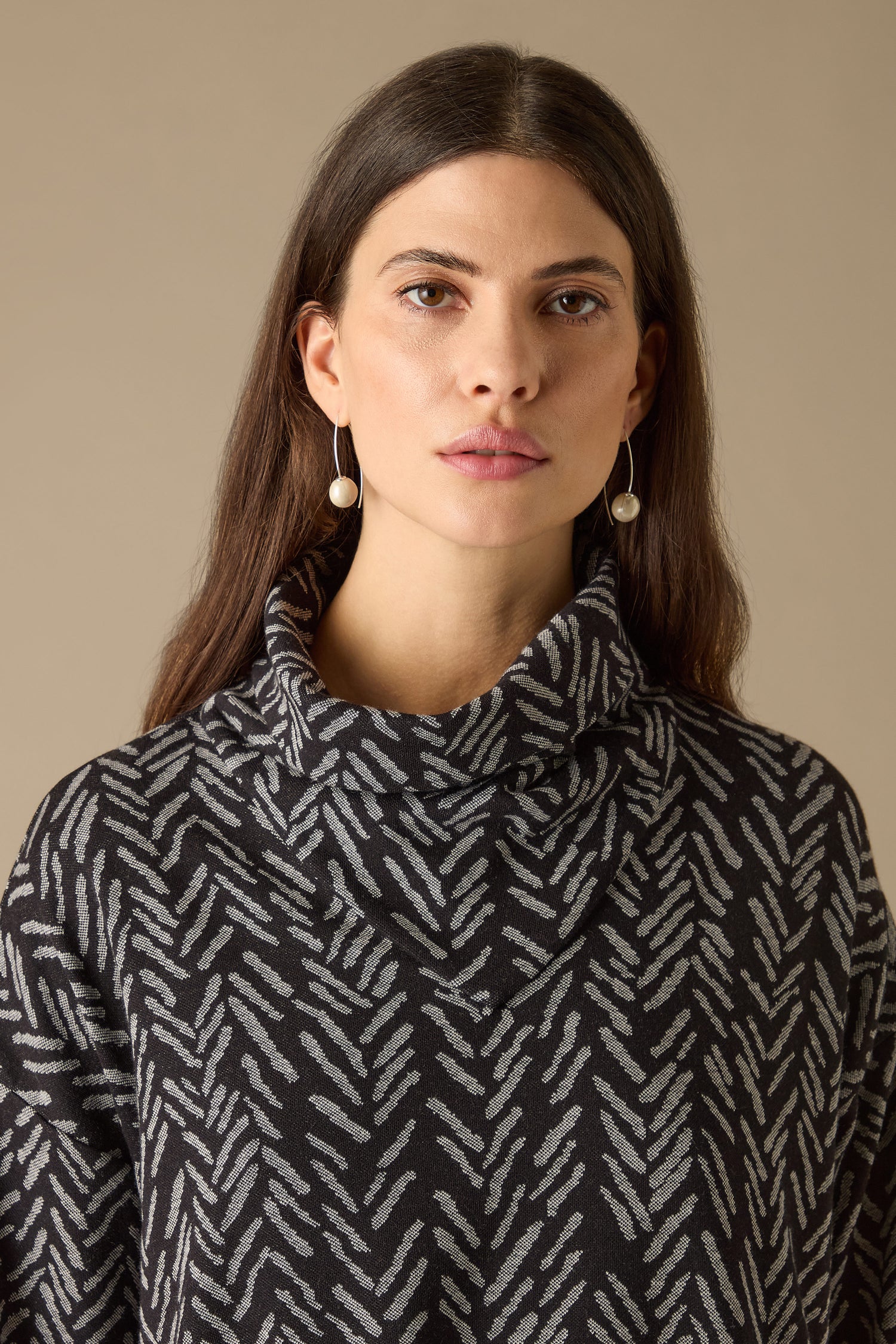 Against a plain backdrop, a long-haired individual models the Herringbone Jersey Jacquard Top, known for its boxy fit and patterned design, accessorized with pearl earrings.