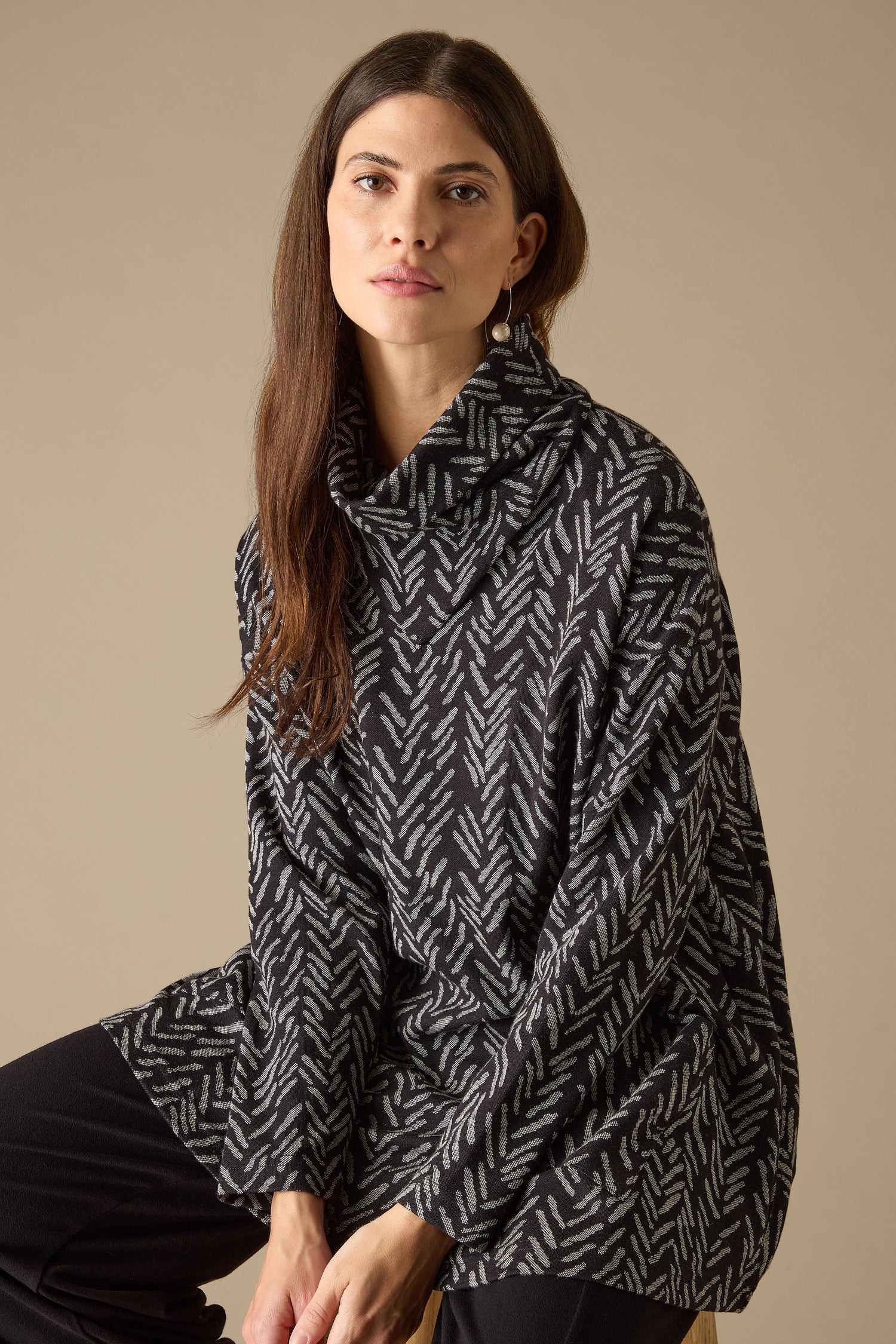 A person with long brown hair models the Herringbone Jersey Jacquard Top, characterized by its boxy fit and funnel neck, while seated on a stool against a plain background.