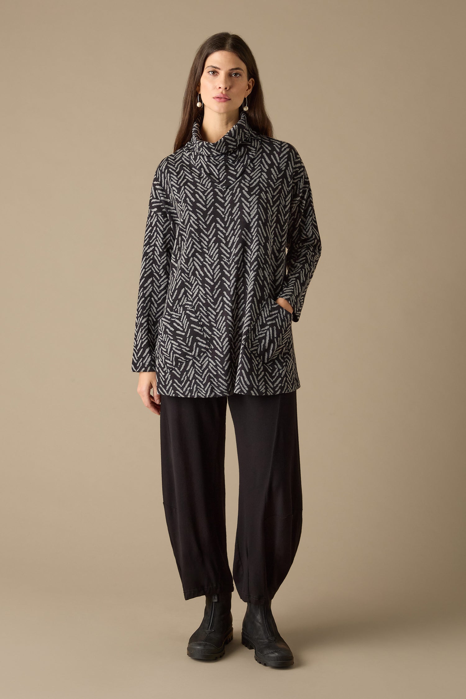 A person stands against a neutral background wearing a Herringbone Jersey Jacquard Top with a stylish boxy fit, paired with black pants and black boots.