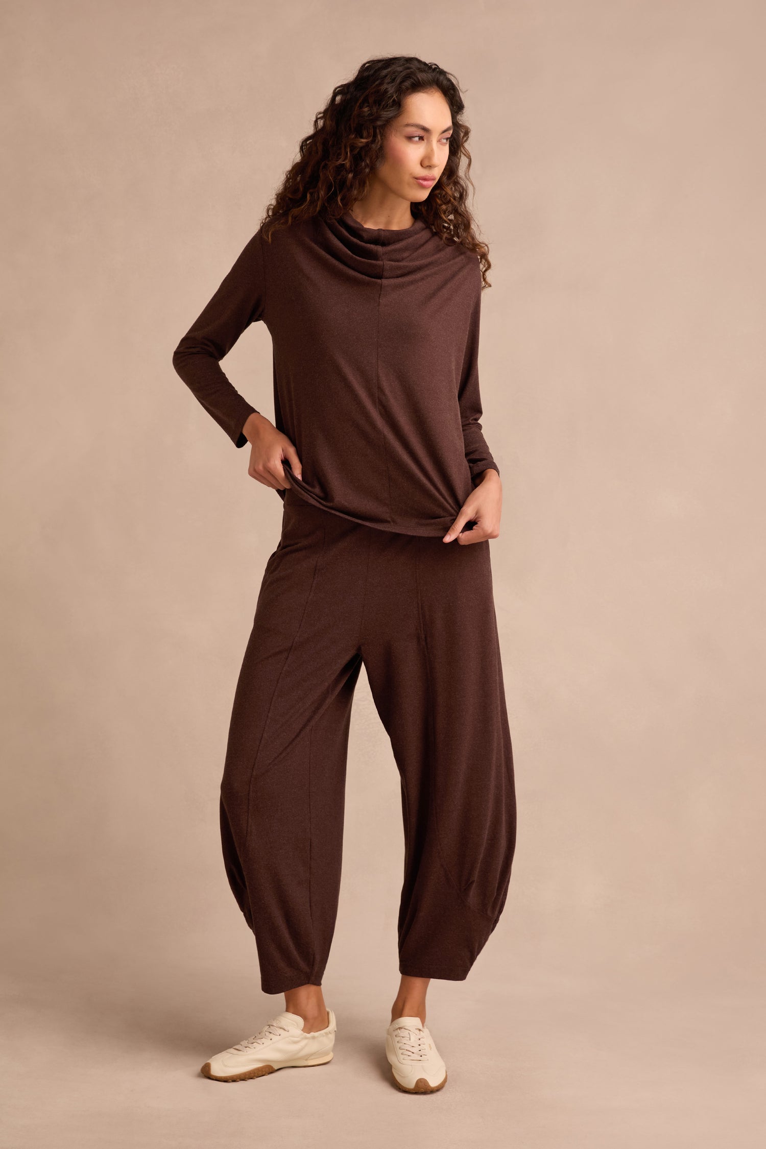 A woman stands against a beige background wearing the Soft Viscose Jersey Ruched Top in brown, paired with matching wide-leg pants and white sneakers.