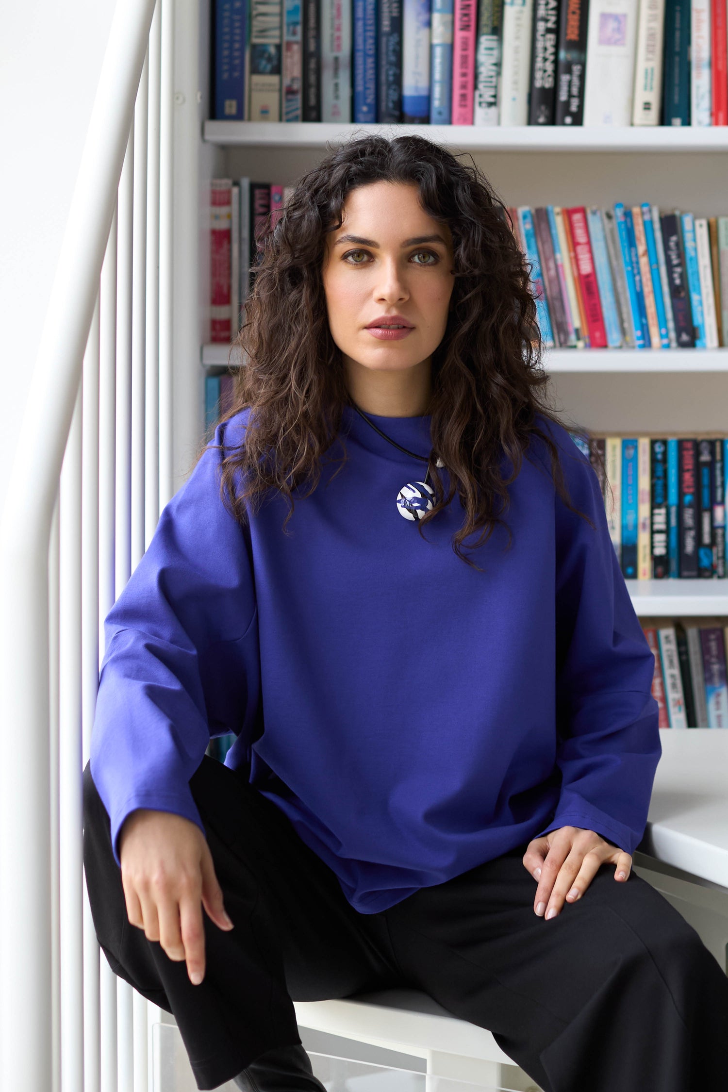 A person with curly hair sits on a chair in front of a bookshelf, exuding effortless style in their blue Ponte Boxy Top and black pants.