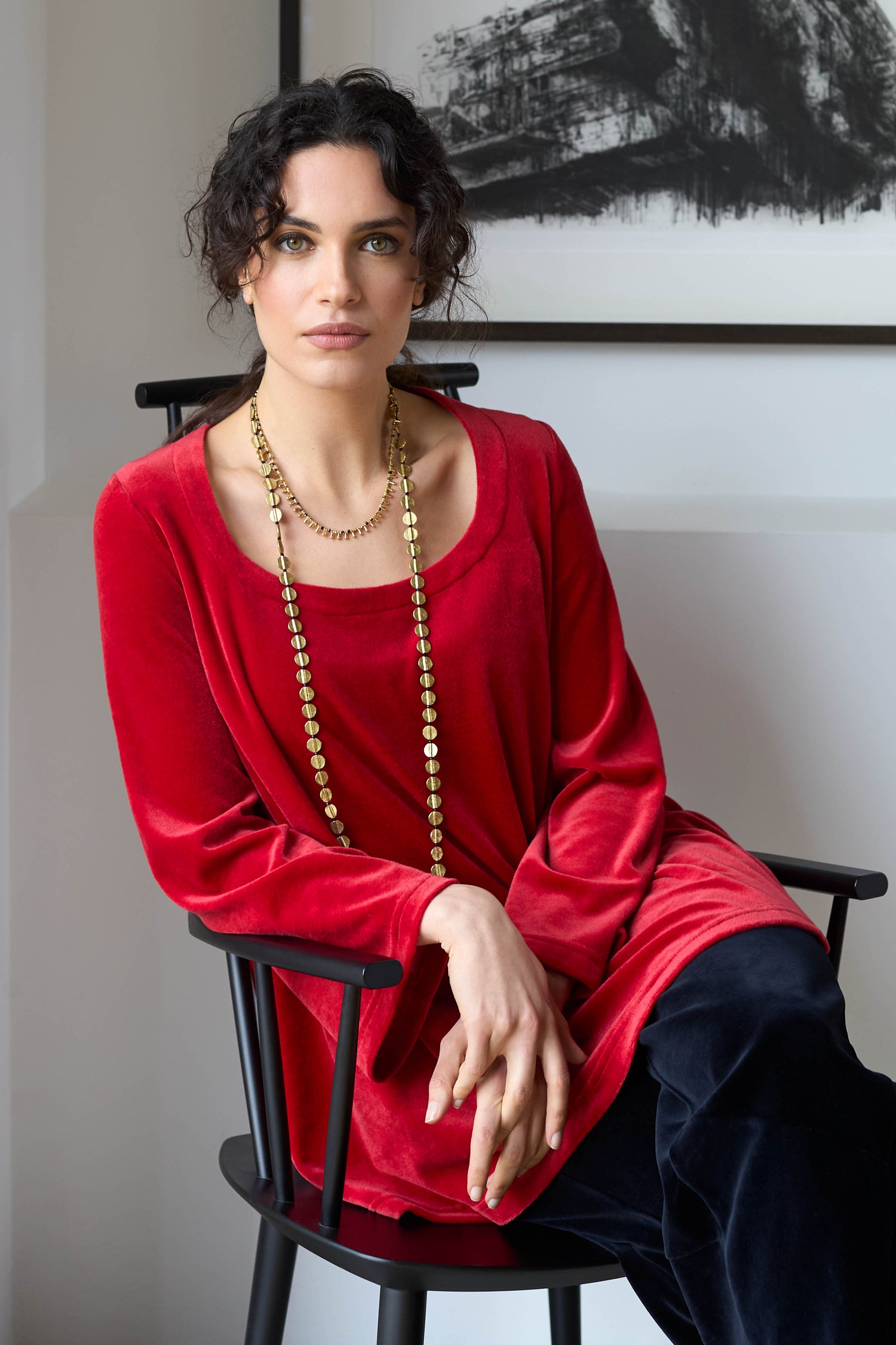 A person with curly hair, wearing a red Velvet Jersey Horseshoe Neck Top and a long necklace, sits on a black chair in front of a painting.