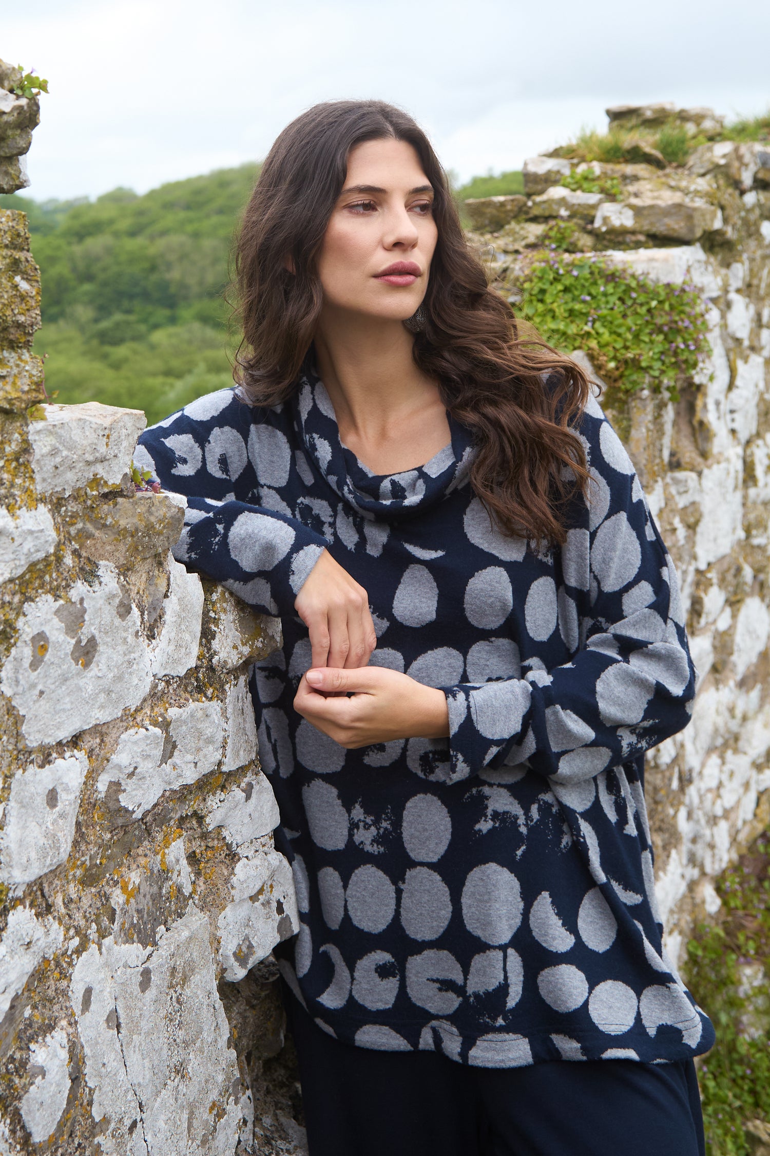 Woman with long brown hair in a Brushed Jersey Spotted Boxy Top (navy blue and gray) stands by a mossy stone wall, gazing into the distance with greenery in the background.