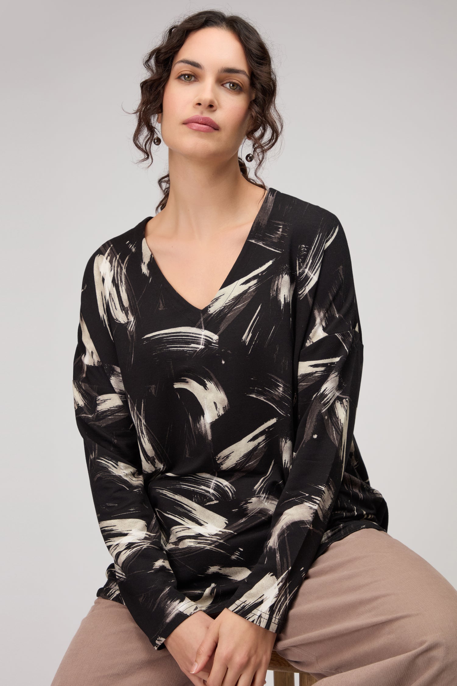 A woman with dark wavy hair wears the Painted Brushstrokes Jersey Top in black and white, paired with beige pants, as she sits on a stool against a plain background.