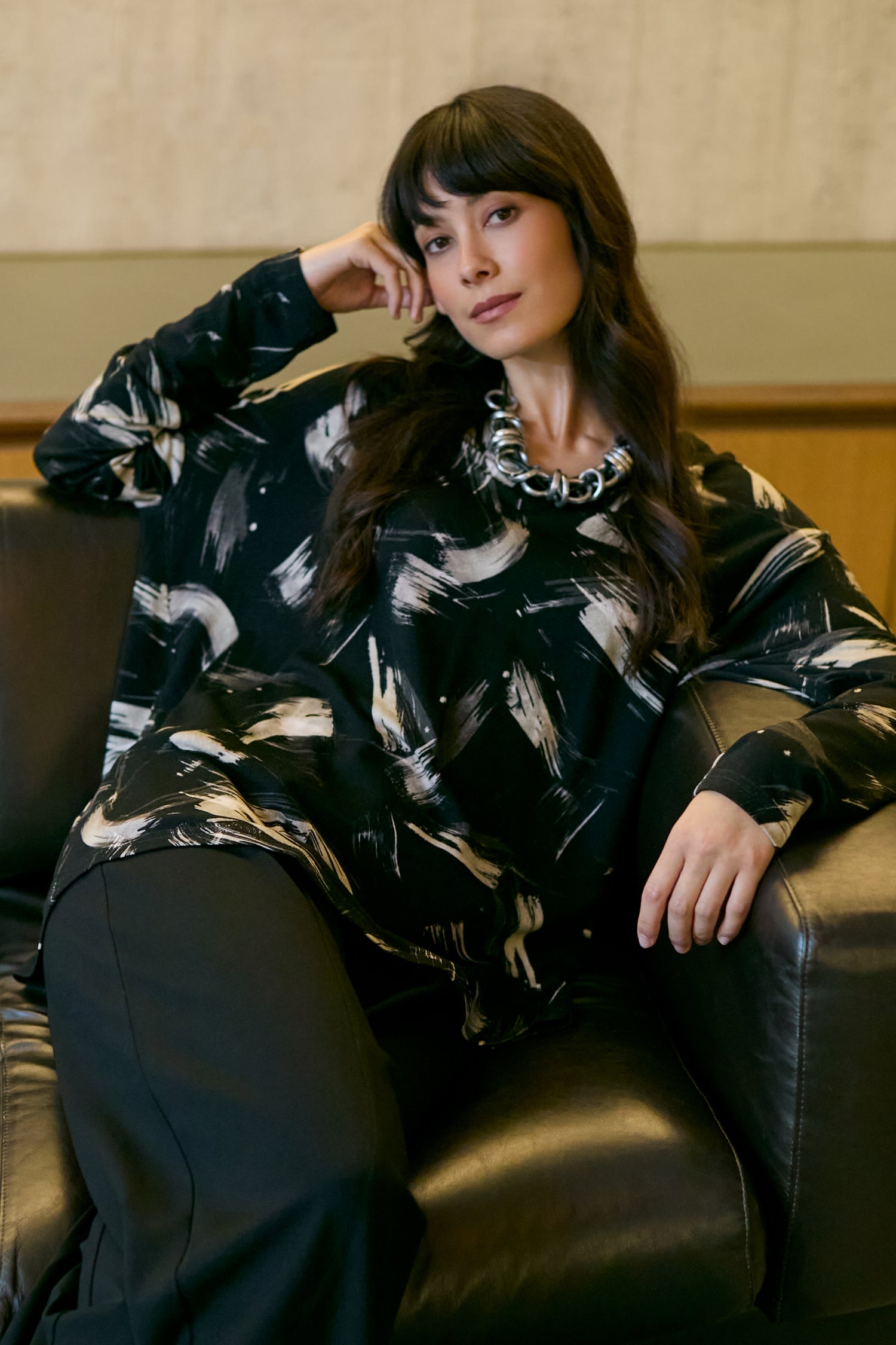 A woman with long dark hair sits on a black leather chair, wearing the Painted Brushstrokes Jersey Top in black and white, paired with black pants and a chunky silver necklace.