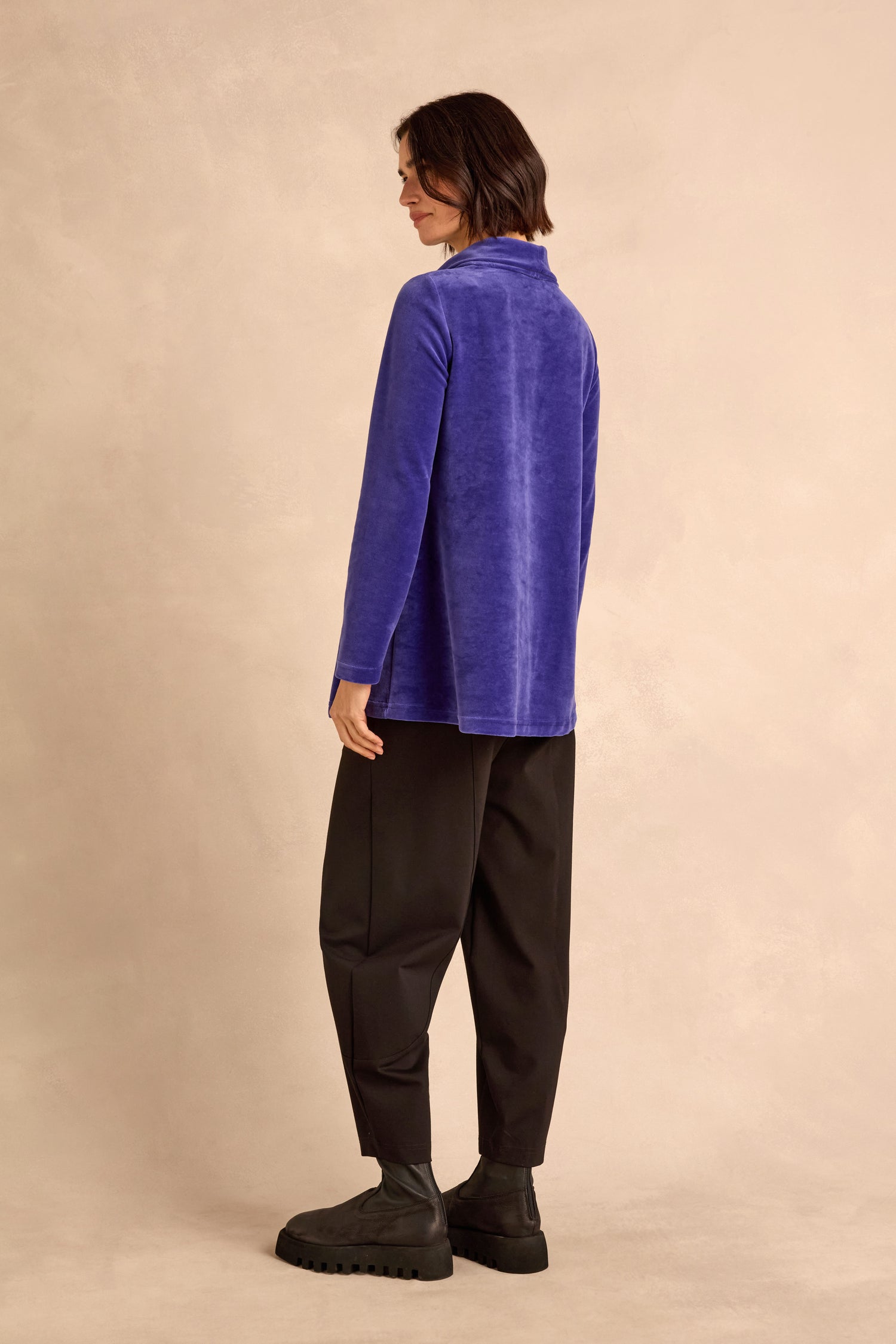 A person stands sideways against a beige background, wearing the Velvet Jersey Cowl Neck Top in blue, paired with loose black pants and black boots.
