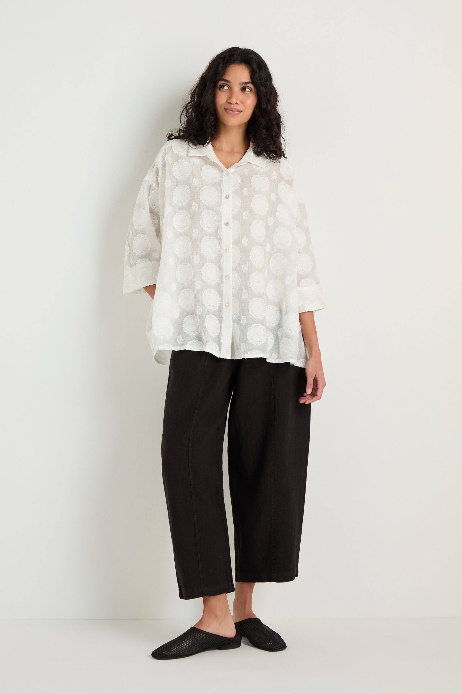 Person stands against a white wall, wearing an oversized Jacquard Circles Shirt in white, black wide-leg pants, and black slip-on shoes.
