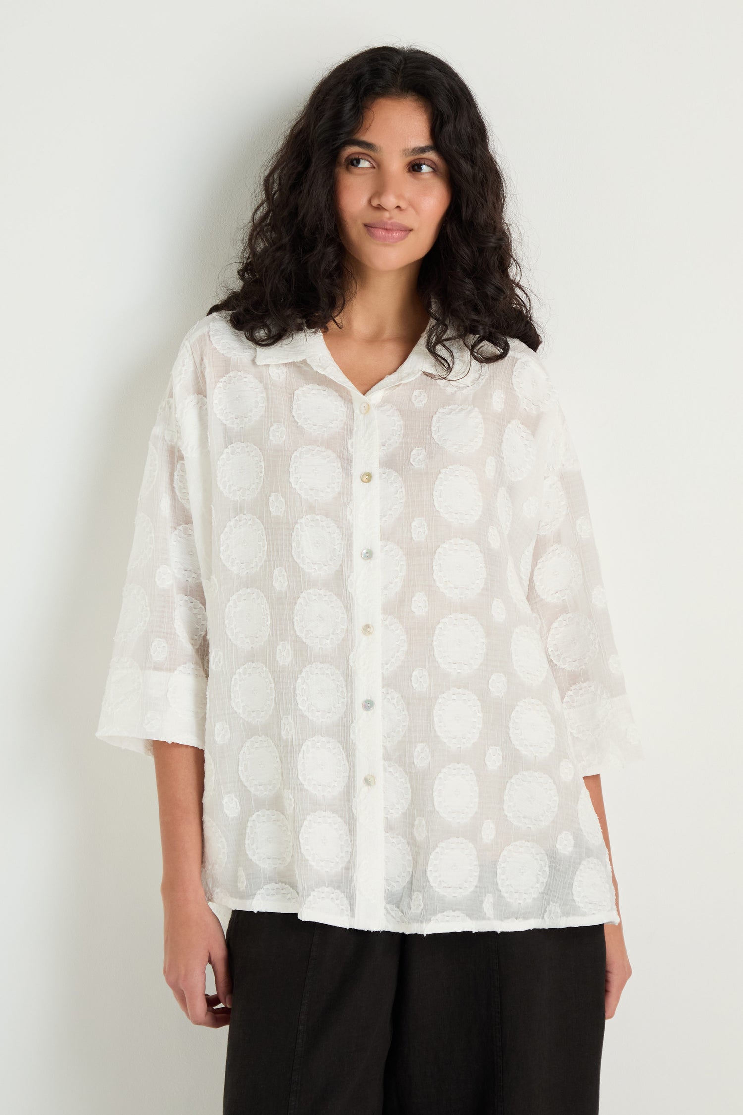 A person stands against a plain light-colored wall wearing the Jacquard Circles Shirt, a loose, white, button-up with textured circle details and an oversized fit.