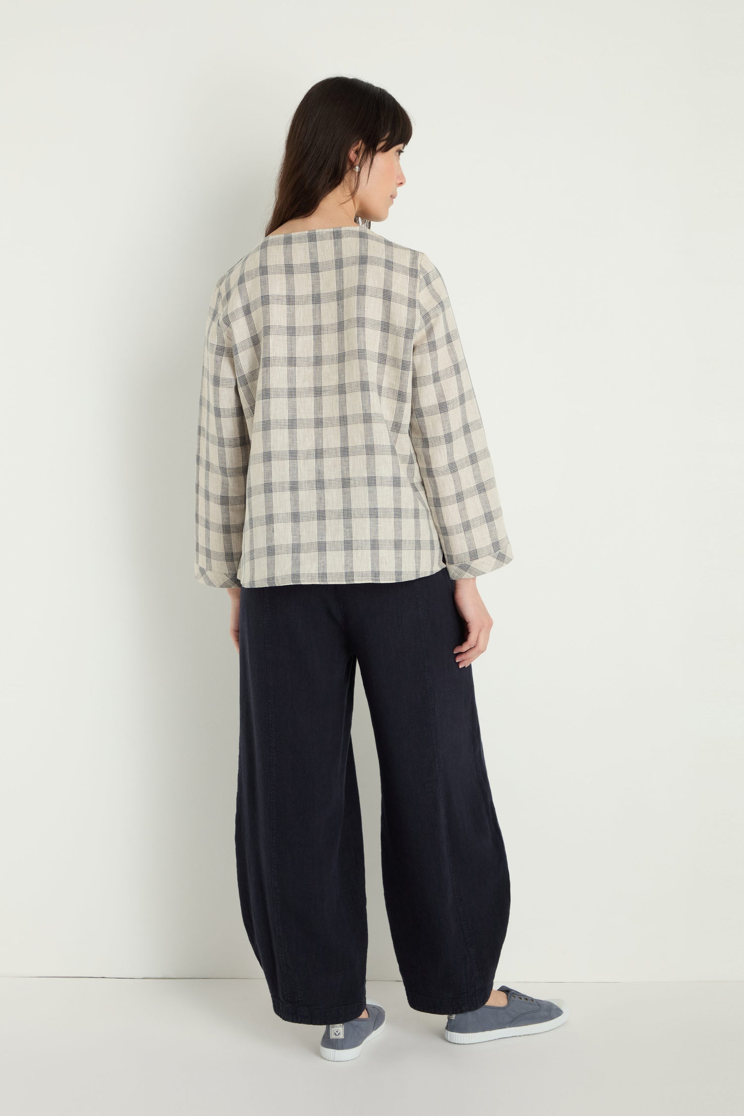 A person stands facing away, wearing the Grid Check Linen Top—relaxed fit beige and gray grid check, paired with wide-leg black pants and gray slip-on shoes against a plain light background.