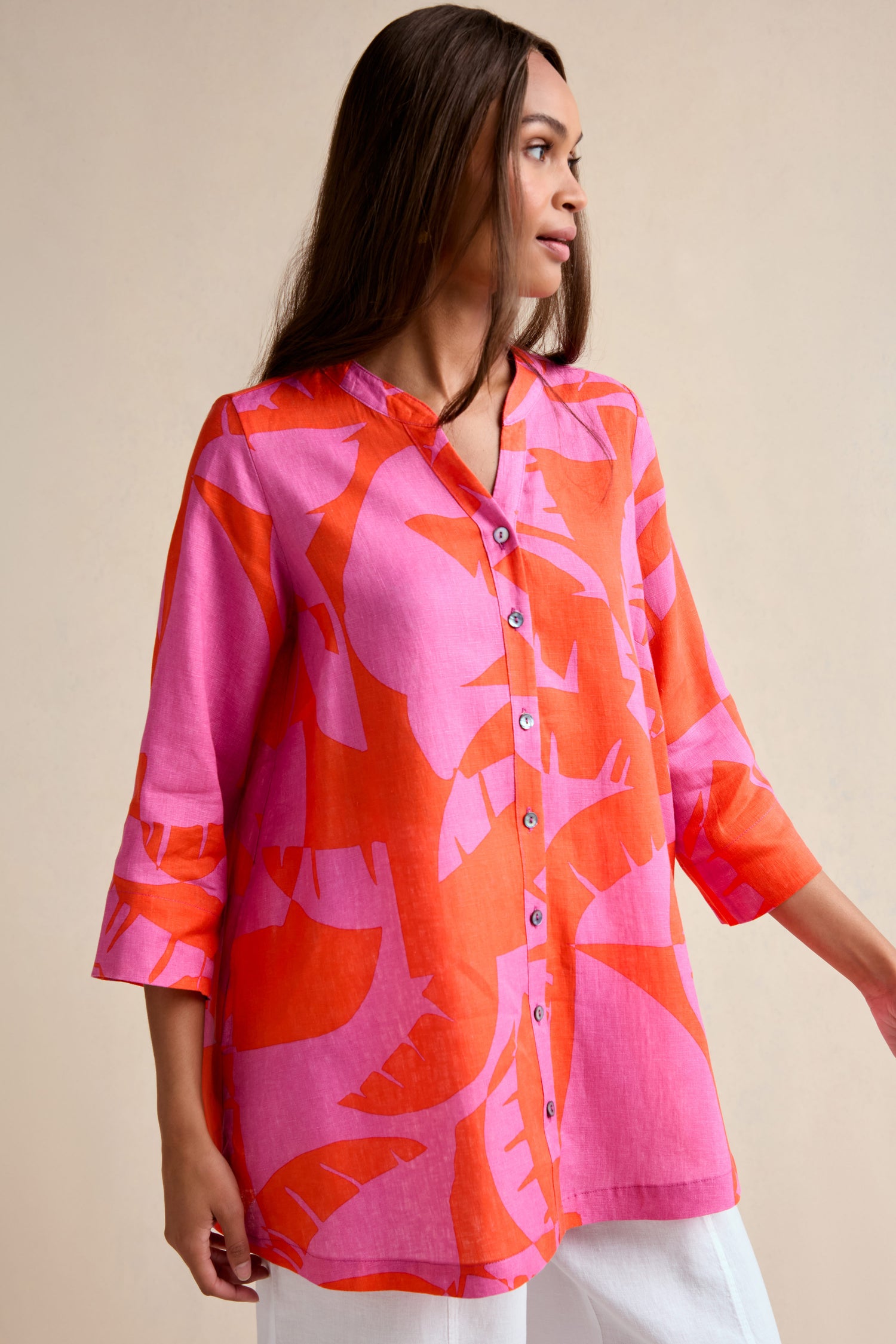 A woman stands against a neutral background wearing the Palm Mirage Linen Shirt, a relaxed-fit, button-up linen shirt featuring a pink and orange abstract pattern.