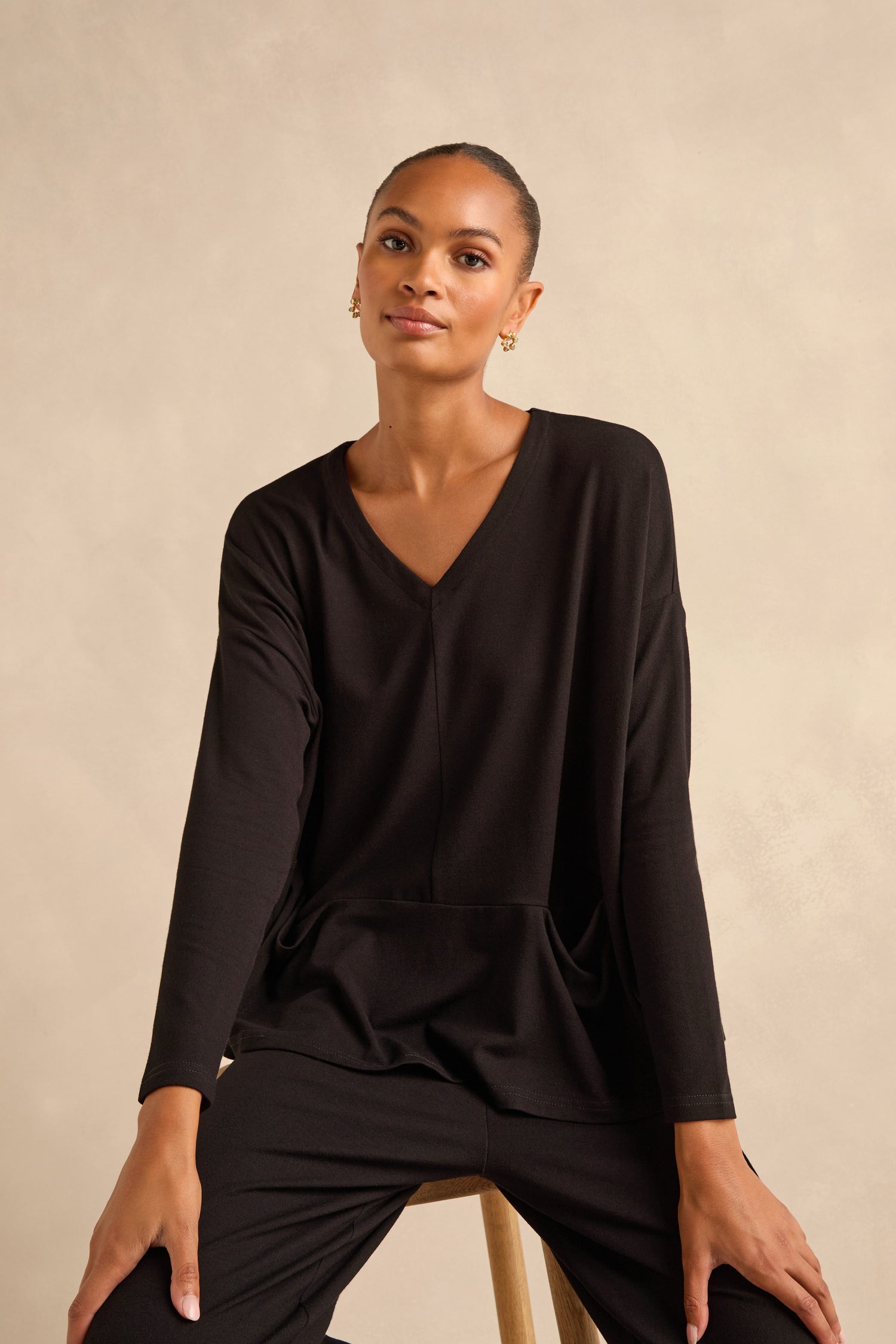 A woman with a neutral expression sits on a stool, wearing the Fluid Crepe Relaxed Top in black with matching black pants against a plain beige background.