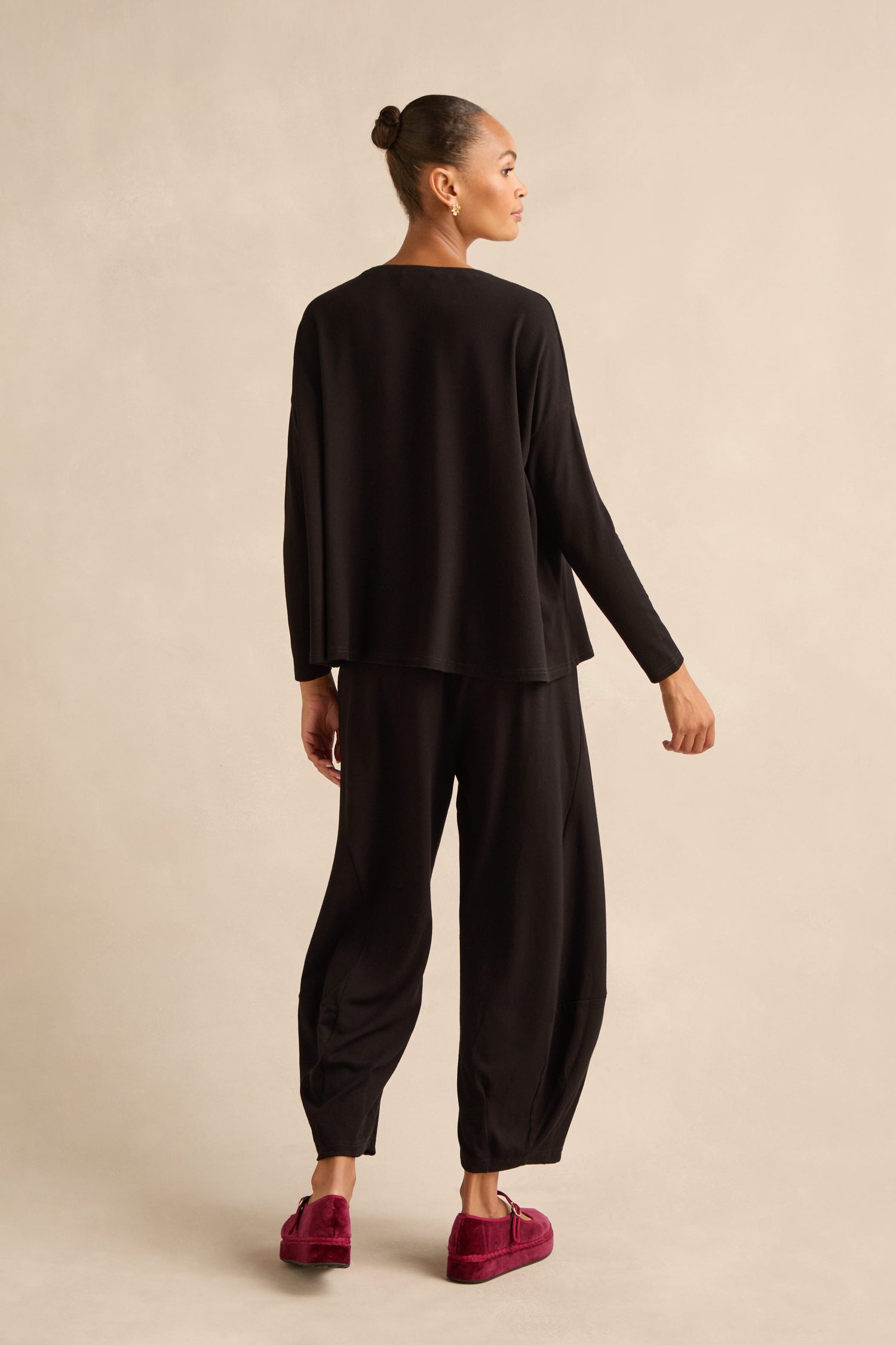 Person standing with back to camera, wearing a loose black Fluid Crepe Relaxed Top with a boxy silhouette and red slippers, against a neutral background.
