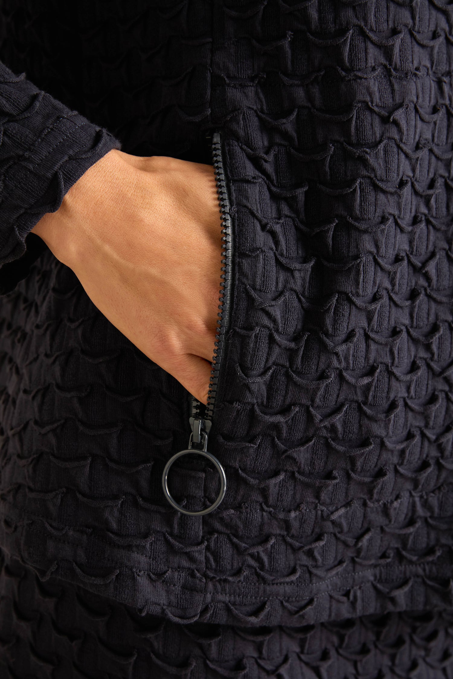 A hand rests inside a zippered pocket on the textured stretch jersey of the dark Origami Pocket Jersey Cowl Neck Top, featuring a circular zipper pull and made in England.