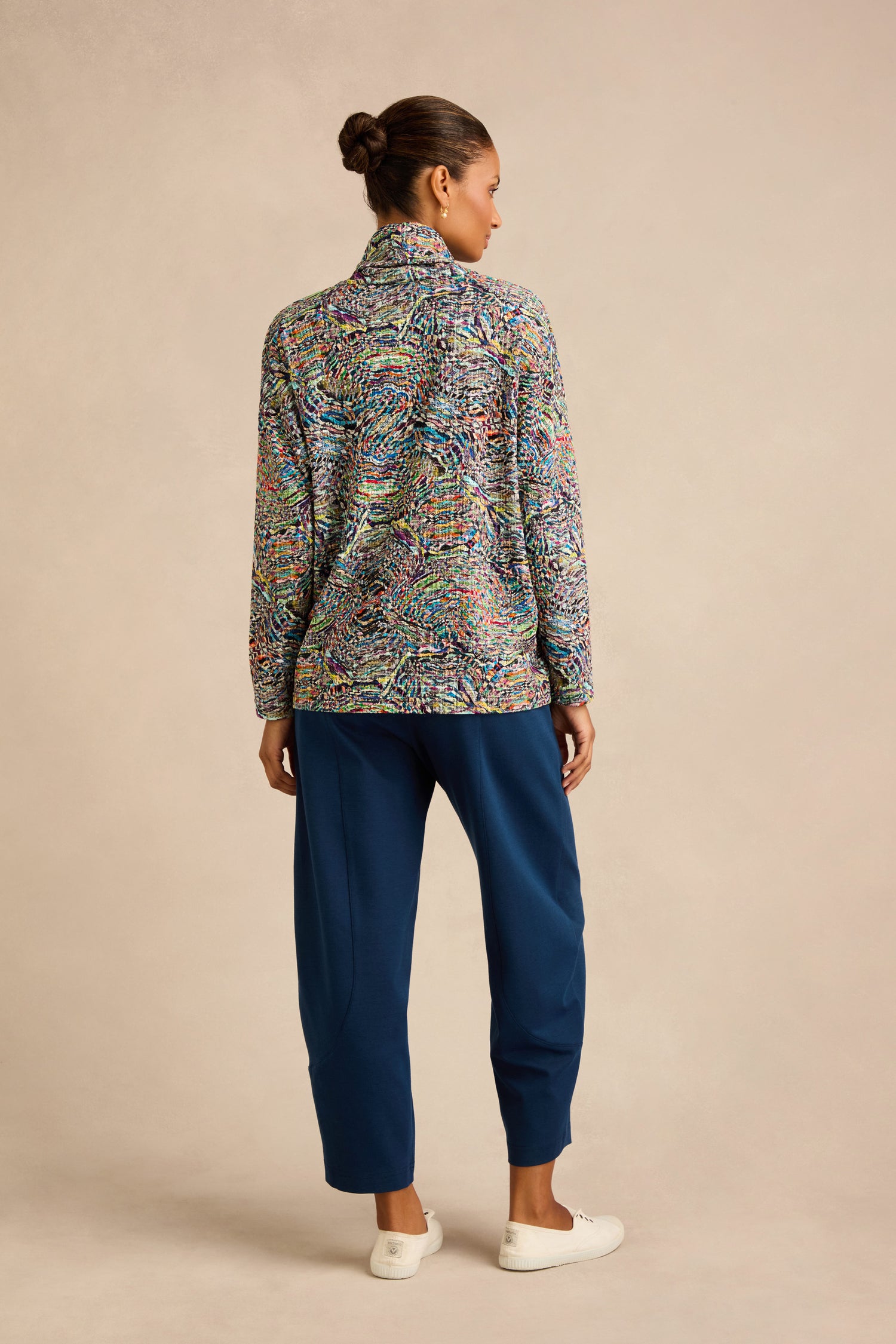 Woman standing with her back to the camera, wearing the Patchwork Fields Jersey Top layered under a colorful patterned jacket, paired with blue pants and white slip-on shoes against a neutral background.