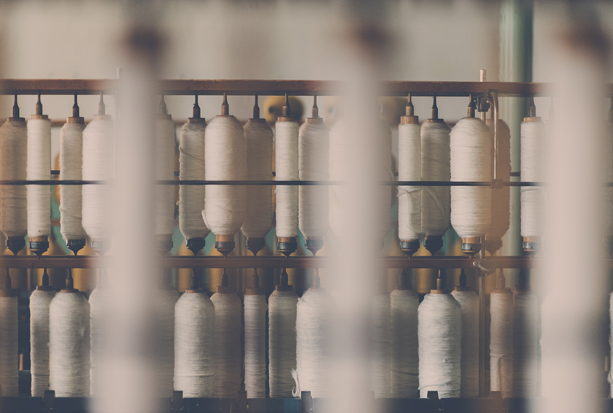 Rows of spindles holding white thread are aligned on a loom in a factory setting.