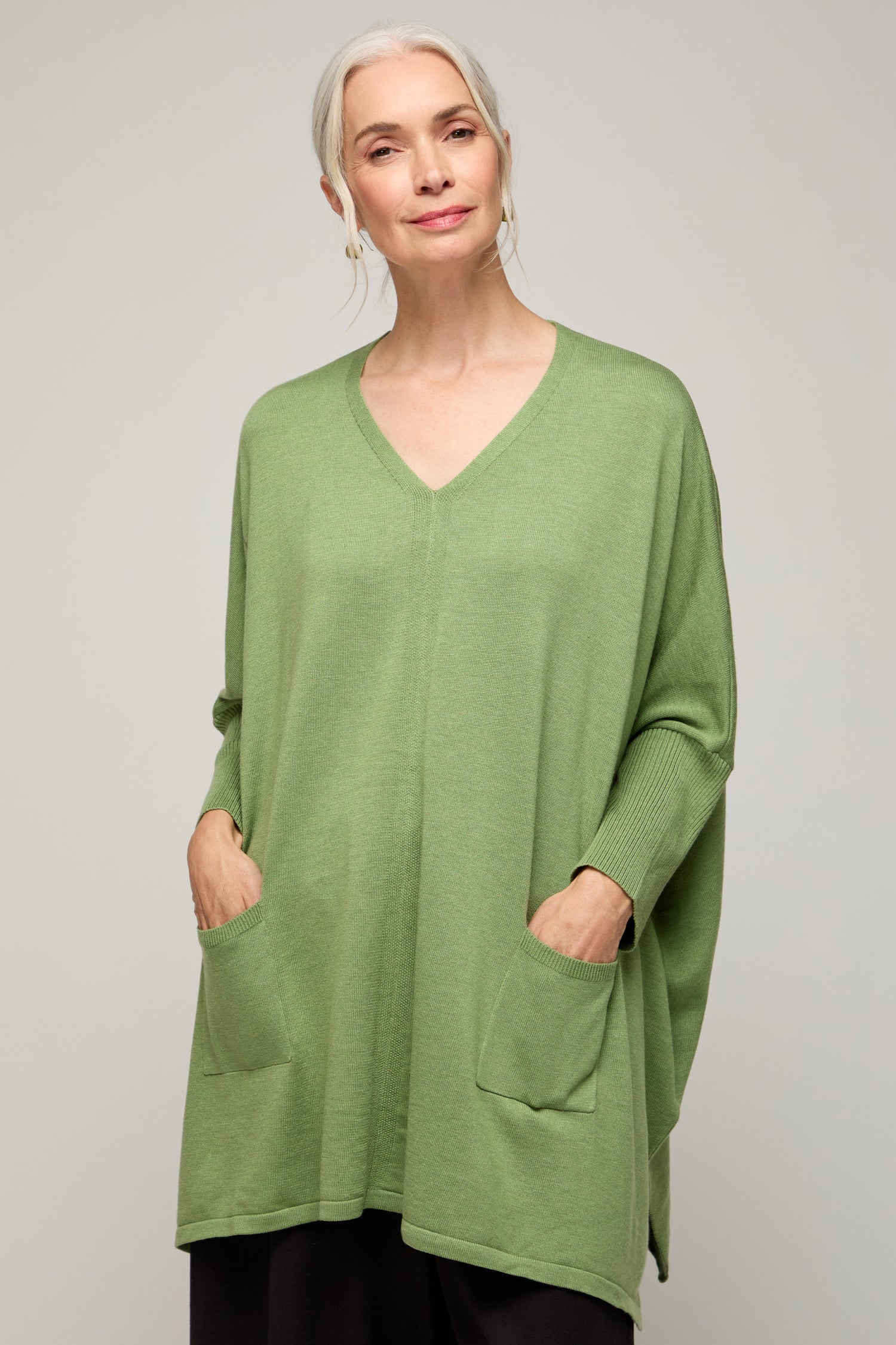 A woman with gray hair models the V Neck Knit—a light green, lightweight sweater featuring front pockets—standing against a plain background for an effortless, minimalist style.