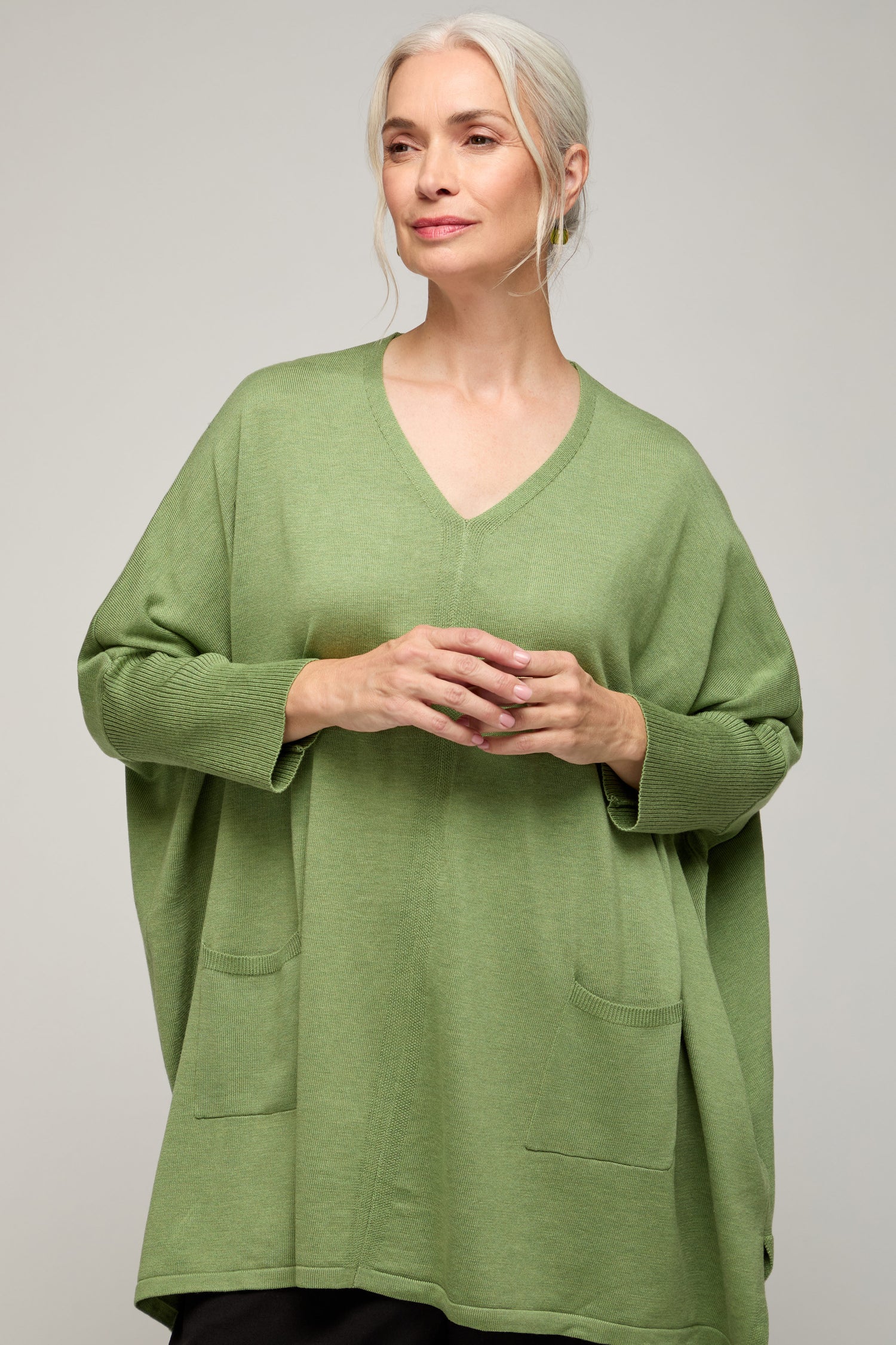 A woman with gray hair wears the V Neck Knit, a loose green sweater made from lightweight fabric with front pockets, embodying minimalist style against a plain light background.