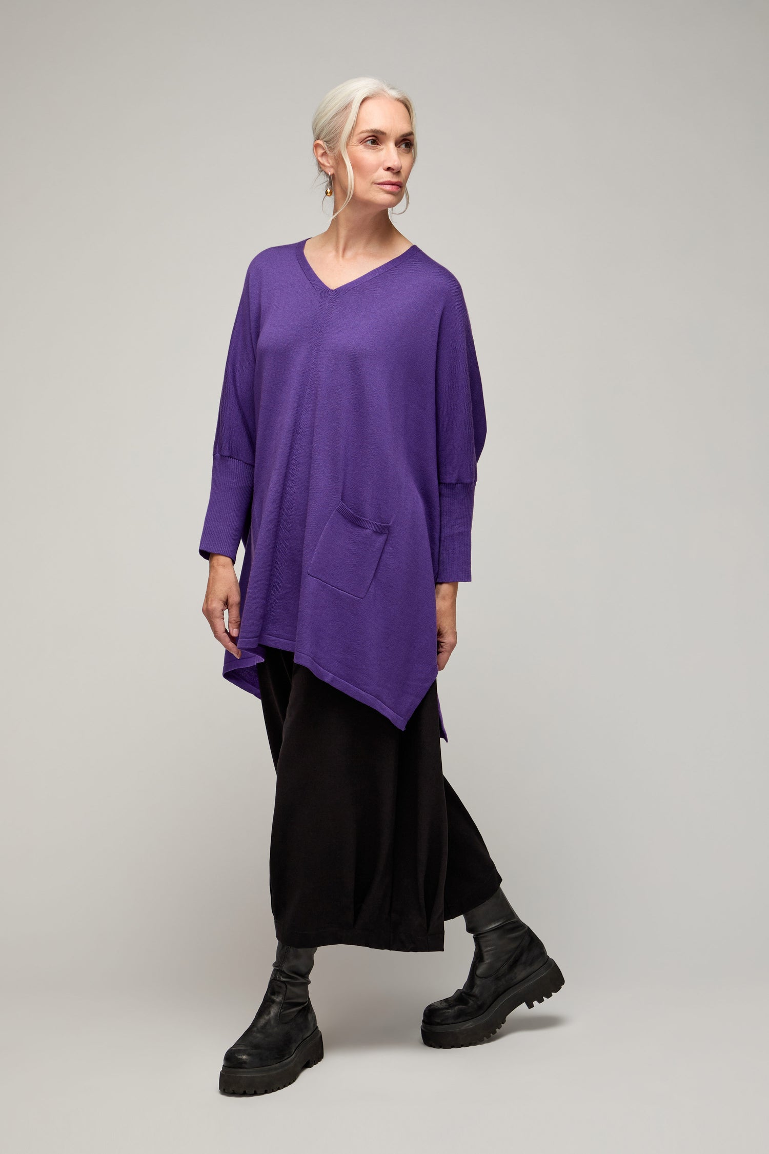 A gray-haired woman wears the V Neck Knit—a loose, lightweight purple top with a front pocket—paired with wide black pants and boots, set against a light background for year-round, minimalist style.
