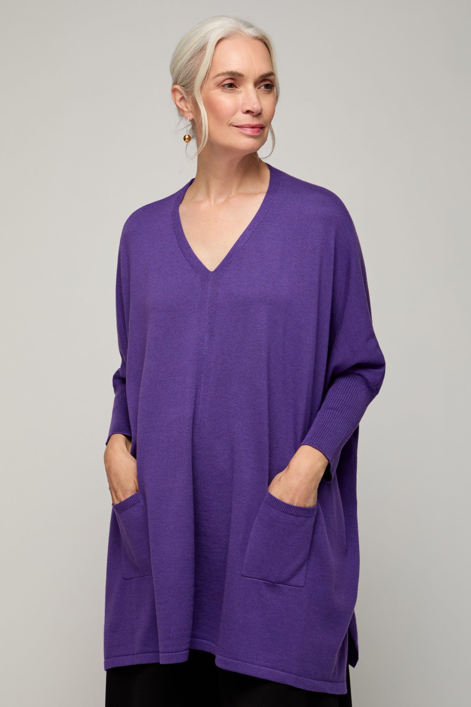 A woman with gray hair models the V Neck Knit, a loose-fitting, lightweight purple sweater with pockets, ideal for year-round wear and shown against a plain light gray background.