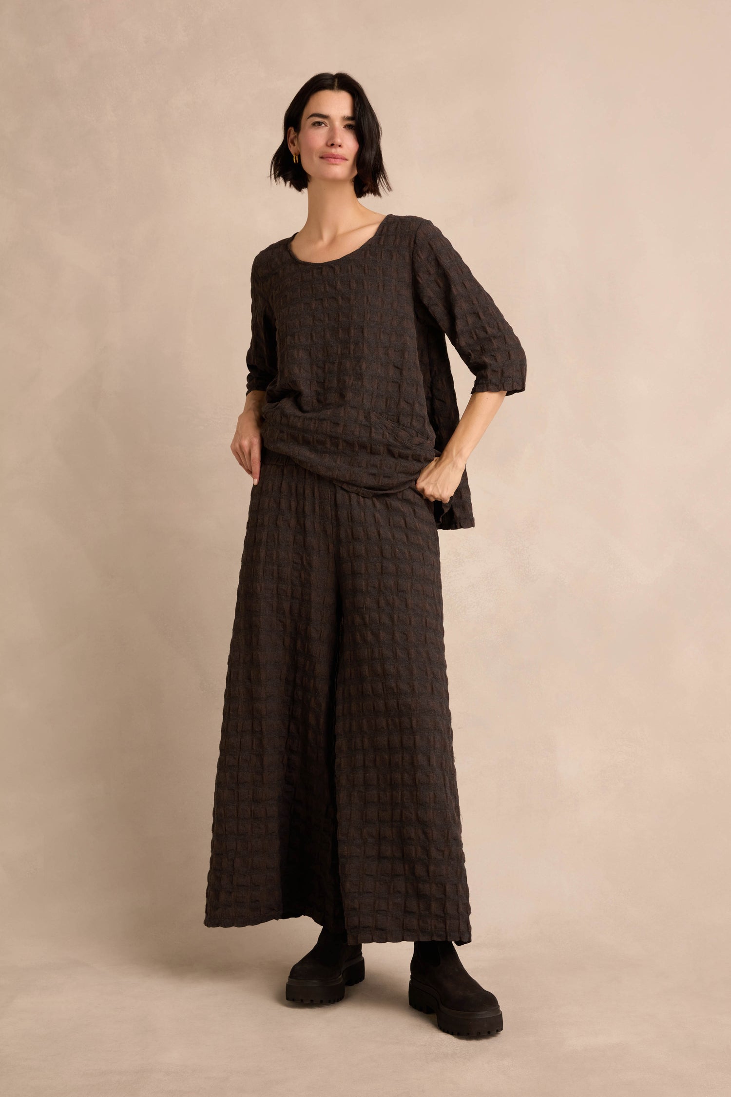 A woman stands against a beige background, wearing the Chocolate Waffle Check Wide Trousers as part of a loose, textured dark two-piece outfit paired with black boots.