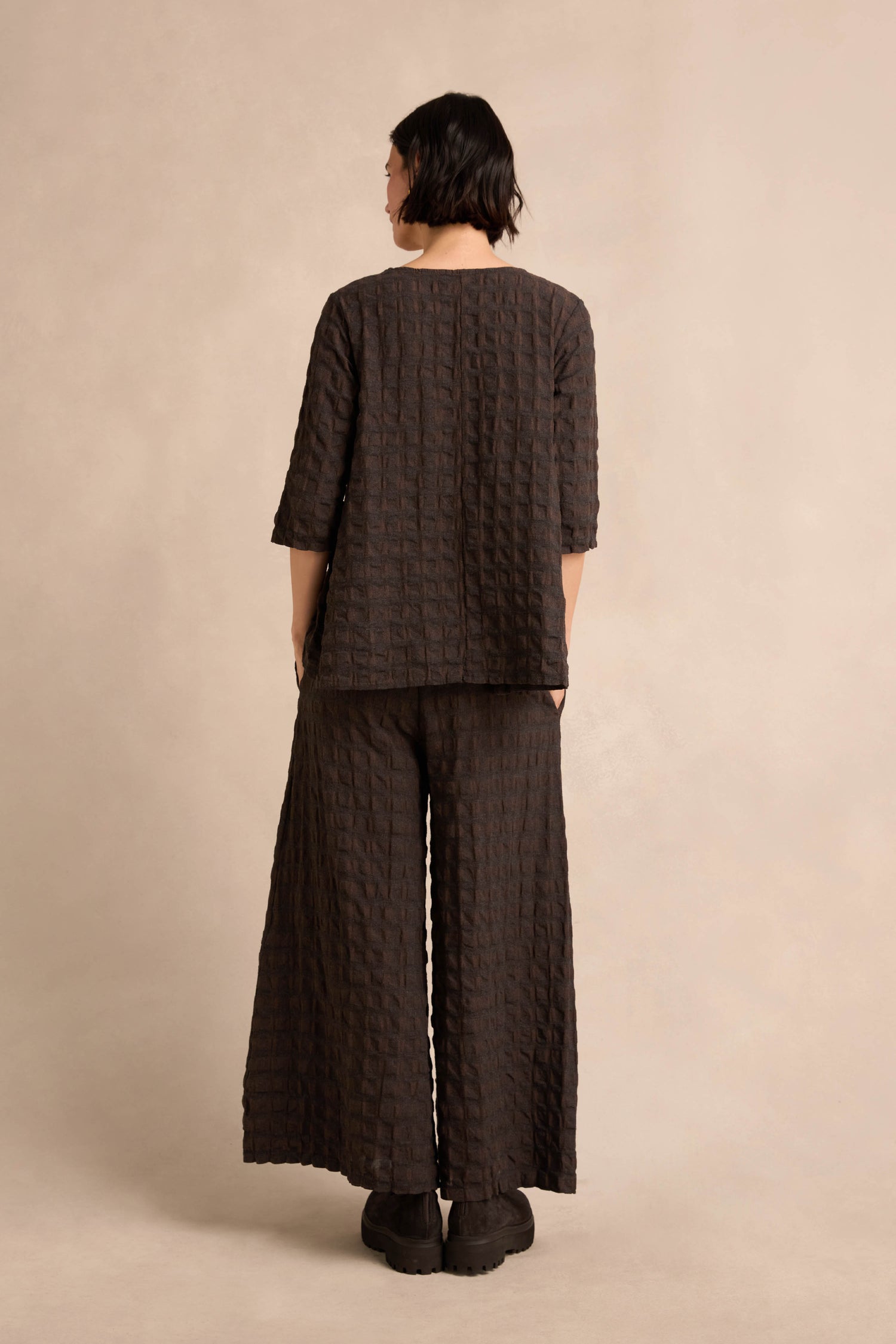 A person stands with their back to the camera in a Chocolate Waffle Check Flared Top, featuring textured dark brown checks and crafted from soft cotton-viscose, paired with wide-legged pants against a neutral background.