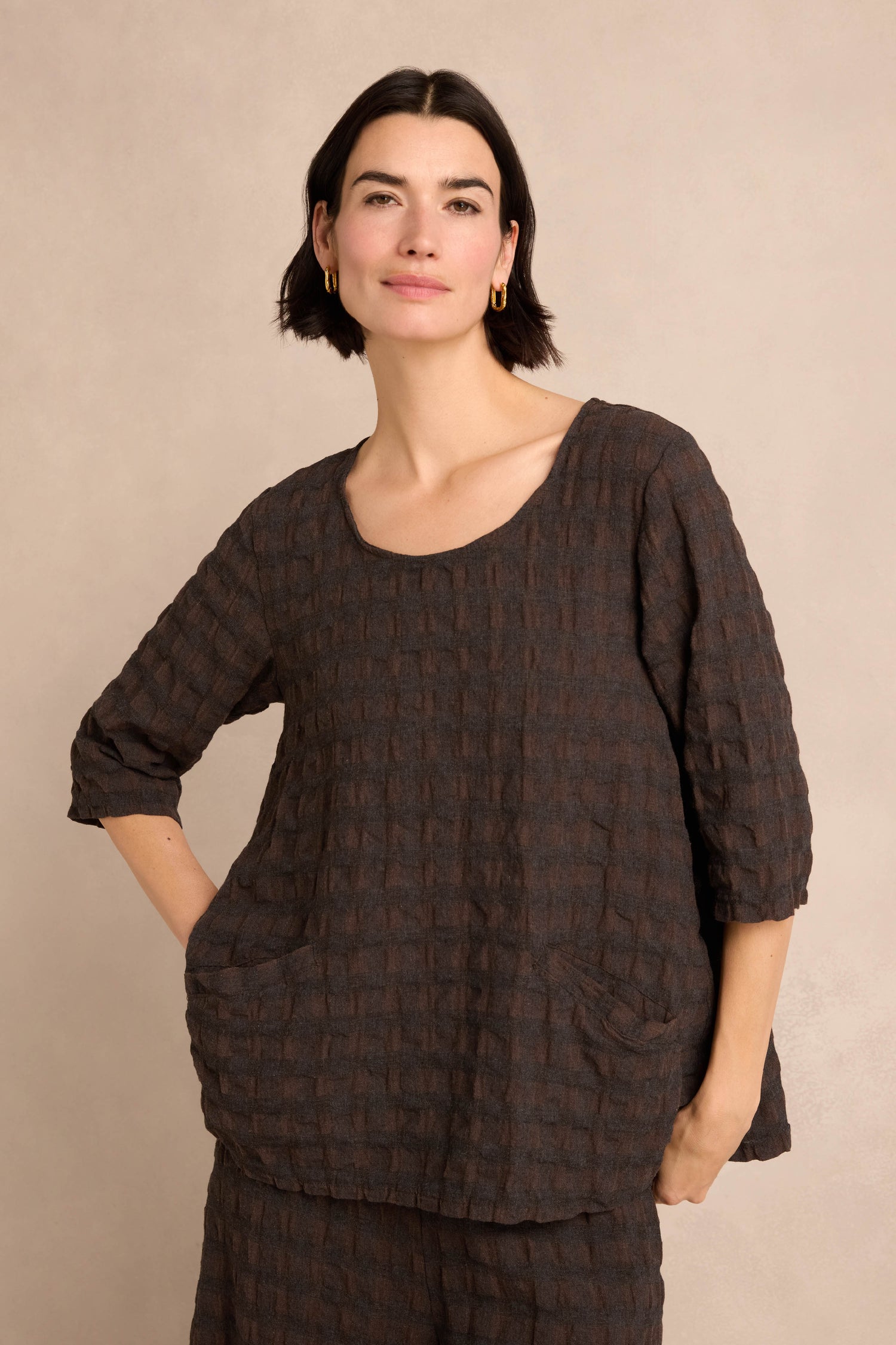 A woman with short dark hair wears the Chocolate Waffle Check Flared Top, featuring three-quarter sleeves, two front pockets, and a loose, textured design crafted from a soft cotton-viscose blend, as she stands against a neutral background.