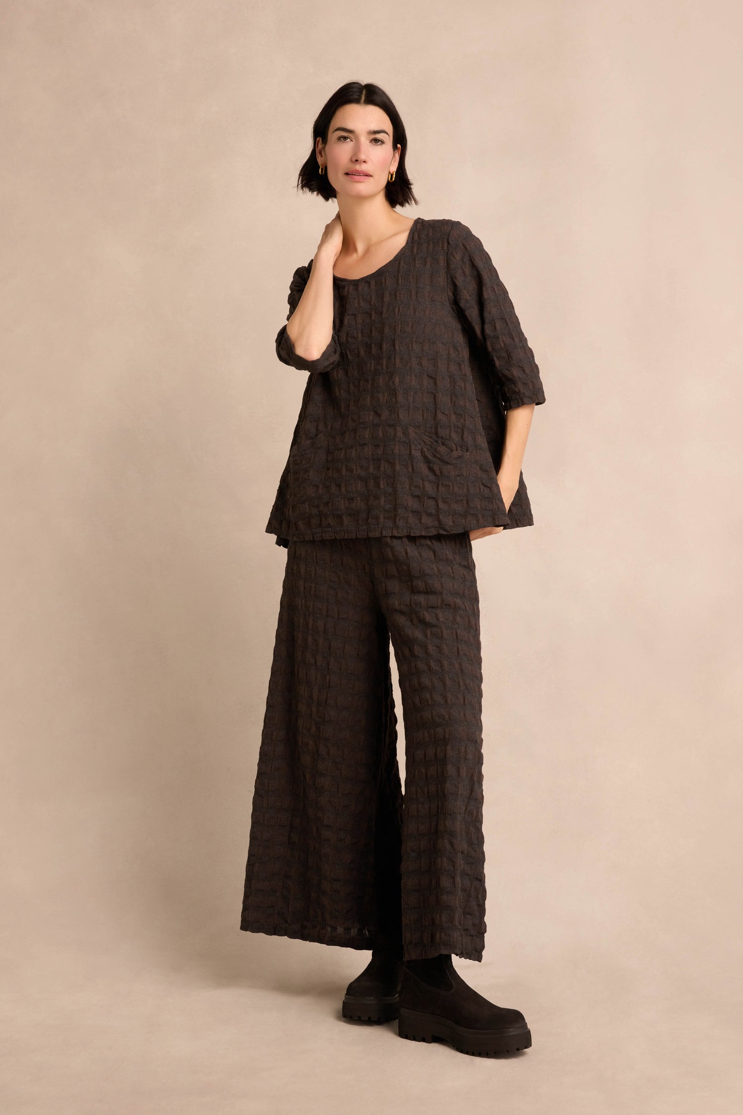 A person stands against a neutral background wearing the Chocolate Waffle Check Flared Top—a loose, textured, dark flared top in cotton-viscose—paired with wide-leg pants and black platform shoes.