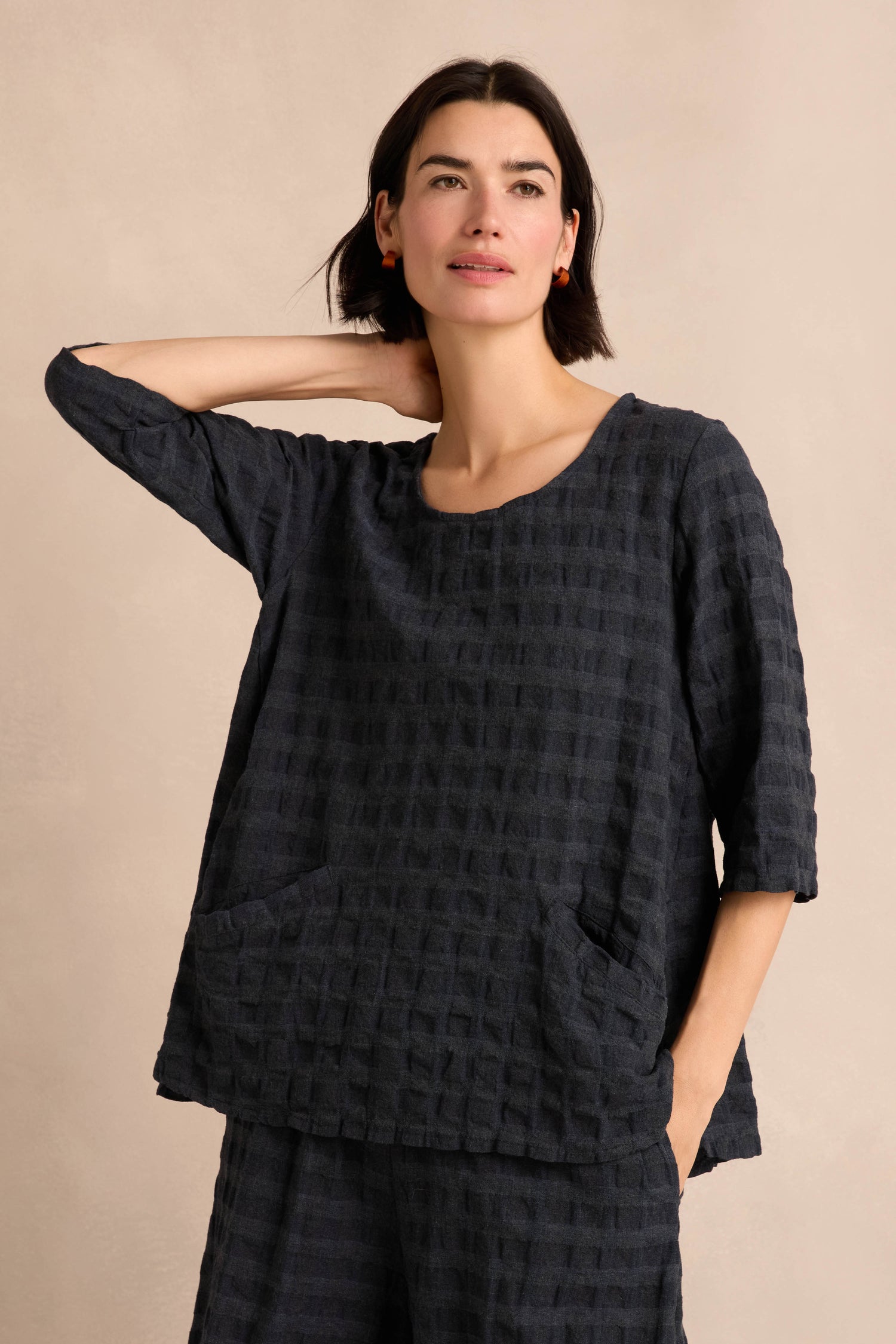 A woman wears the Navy Waffle Check Flared Top, featuring three-quarter sleeves, pockets, and a dark cotton-viscose waffle check pattern, as she stands against a neutral background with one hand raised behind her head.