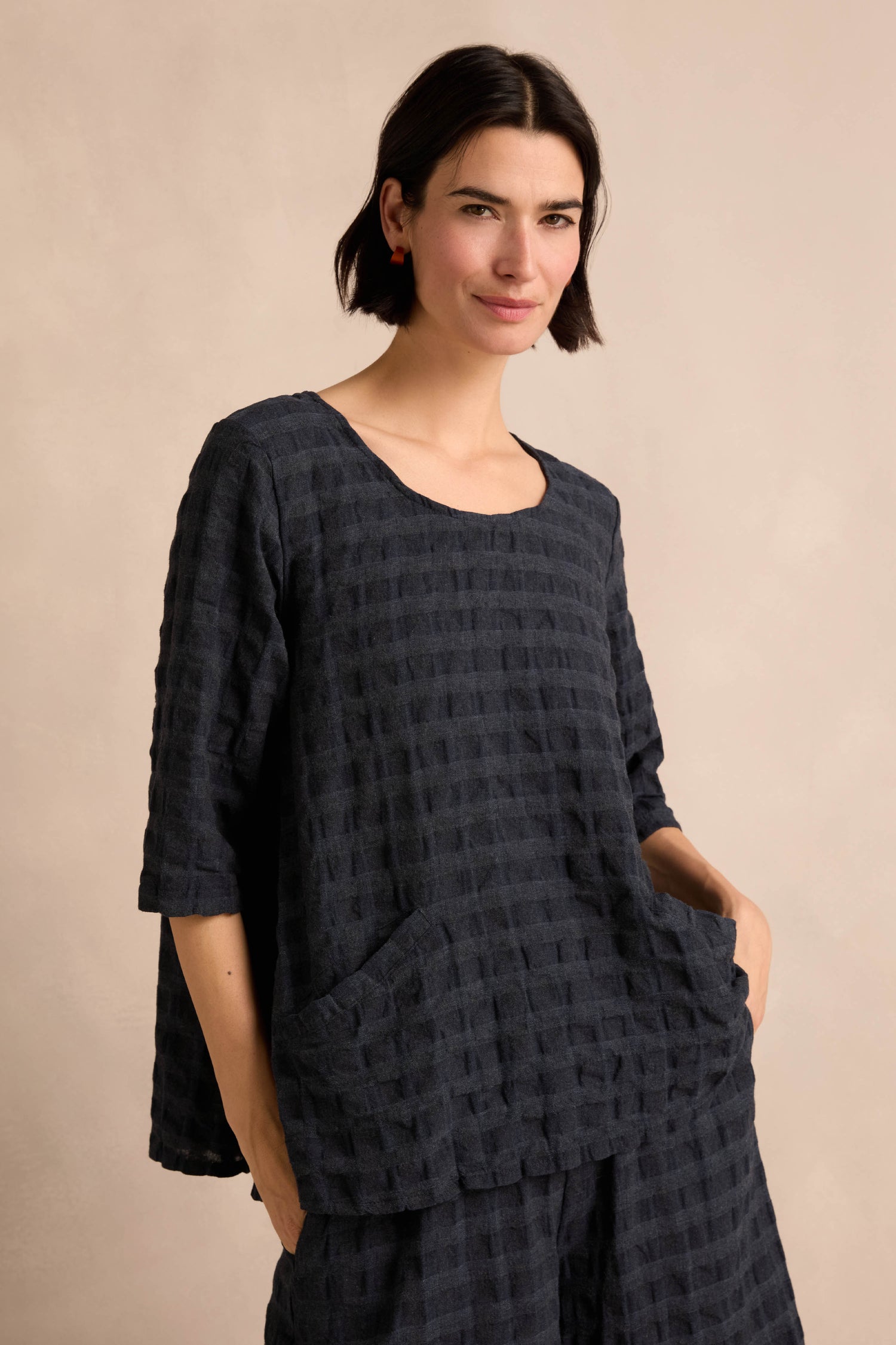 A woman with short dark hair wears the Navy Waffle Check Flared Top, featuring a loose fit with textured pockets, paired with matching pants in a soft cotton-viscose blend, standing against a plain light background.