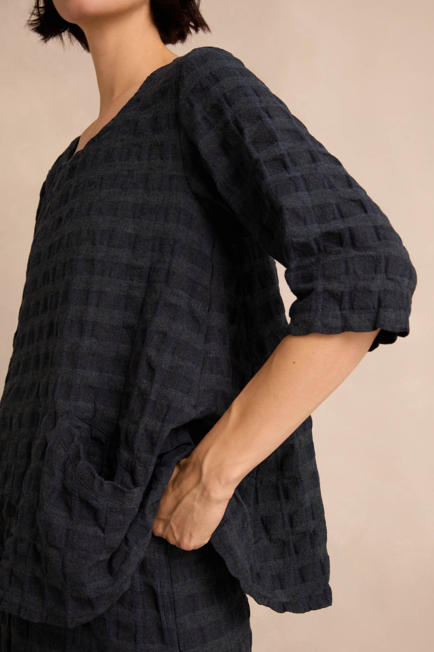 A person wearing the Navy Waffle Check Flared Top, featuring a loose, textured design with three-quarter sleeves, stands with one hand in the pocket against a neutral background.