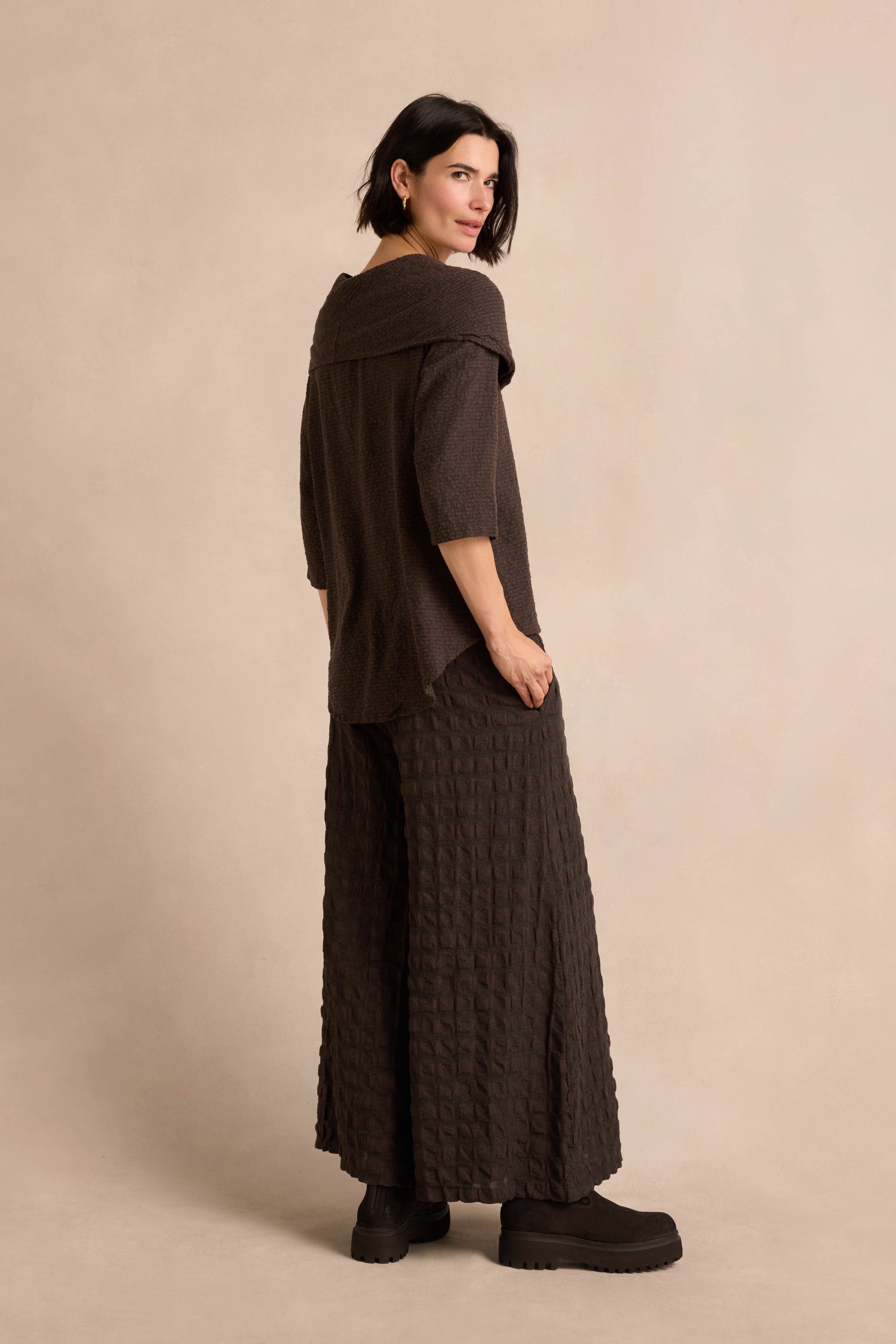 A woman stands sideways against a neutral background, wearing the Chocolate Draped Neck Top—crafted from a loose brown cotton-viscose jersey with wide sleeves and an elegant, asymmetric cowl neckline for added sophistication—paired with matching wide-leg pants and black platform shoes.