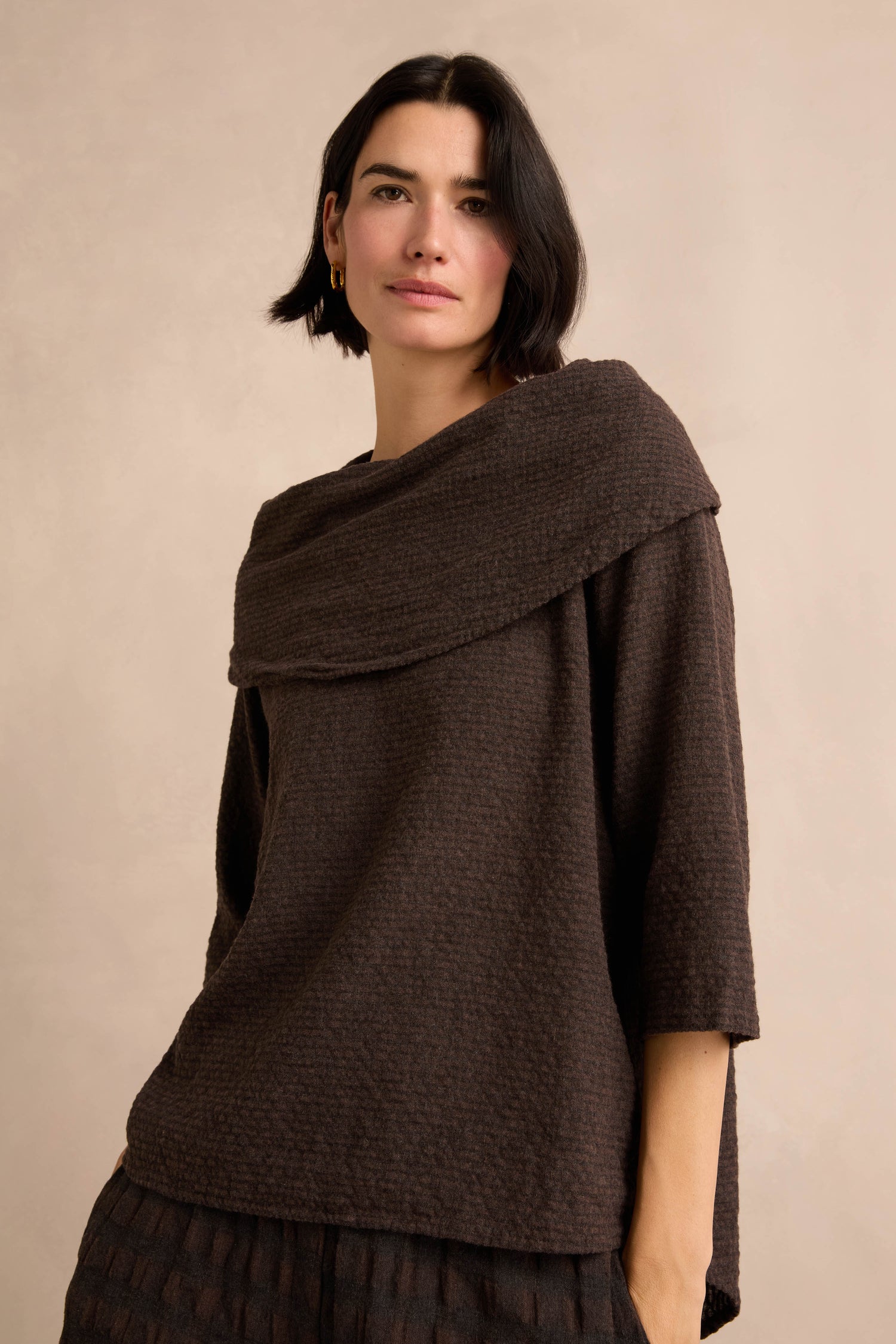 A woman with short dark hair wears the Chocolate Draped Neck Top—crafted from textured brown cotton-viscose jersey and featuring a wide, asymmetric cowl collar—as she stands against a neutral beige background.