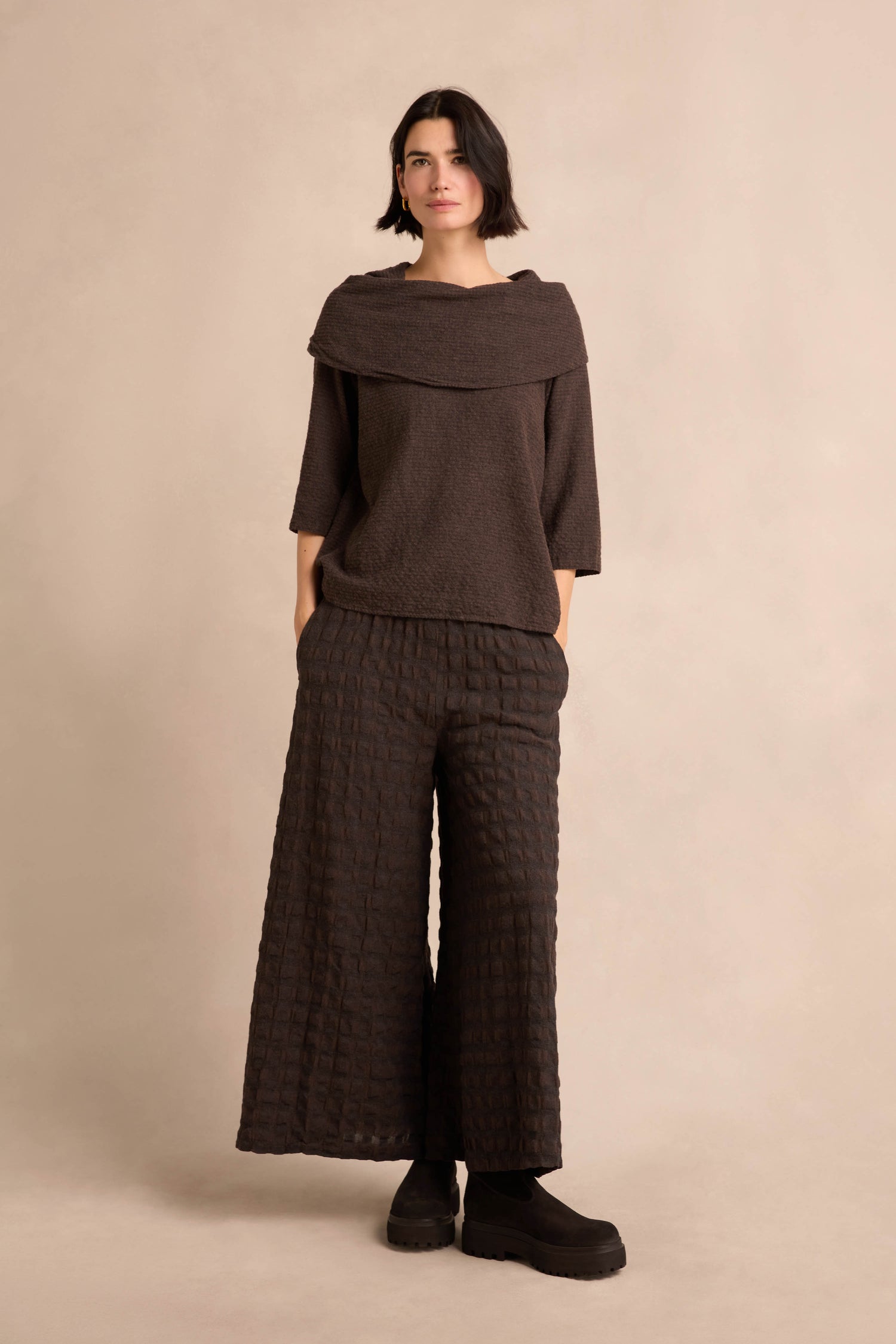 A person stands against a plain background wearing the Chocolate Draped Neck Top, a loose brown piece crafted from cotton-viscose jersey with a wide collar, paired with wide-legged textured brown pants and black shoes.