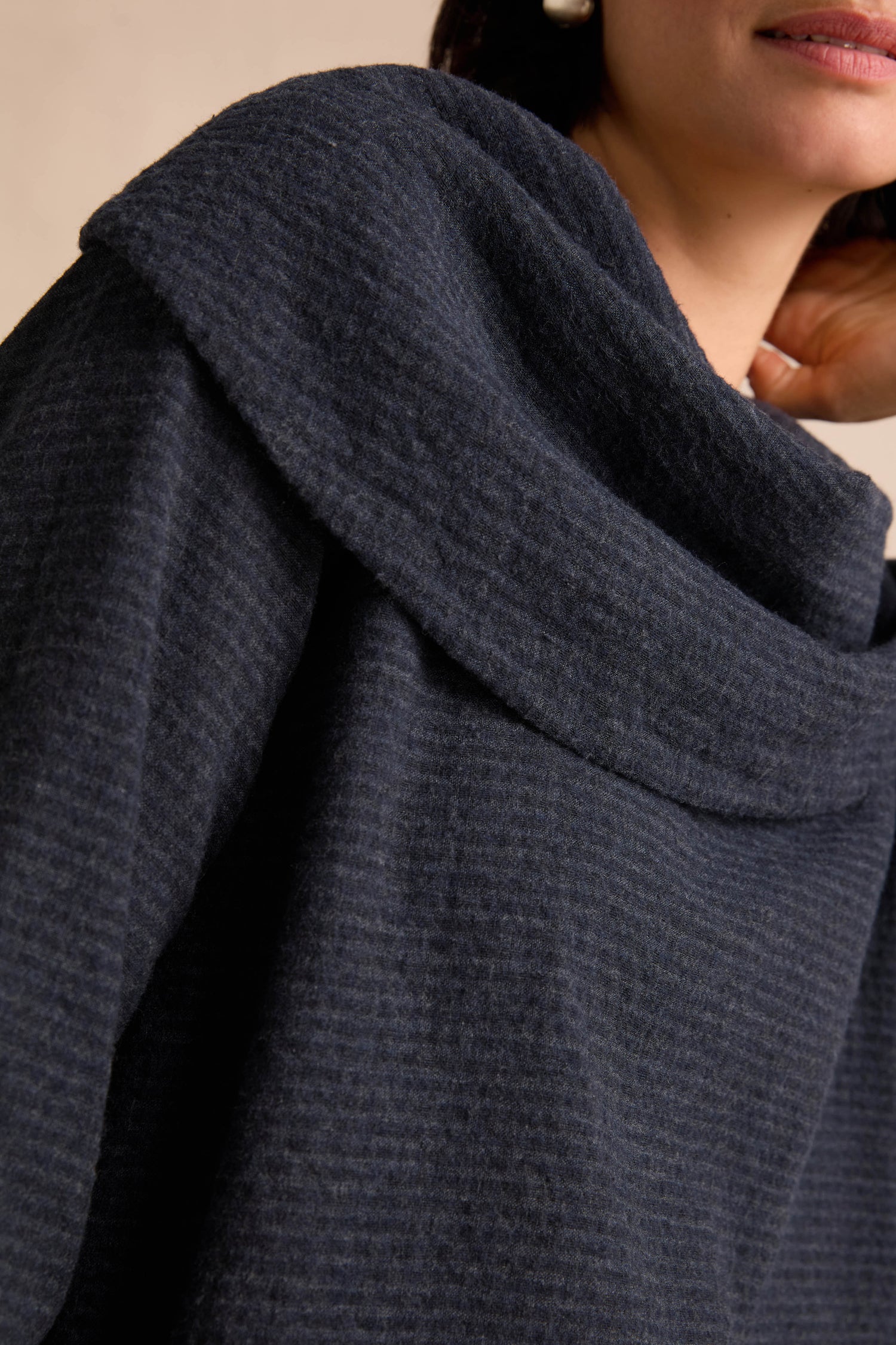 Close-up of a person wearing the Navy Draped Neck Top, featuring a textured dark navy hue and a wide, folded draped collar. The relaxed fit accentuates the soft structure of its cotton-viscose jersey fabric. Part of the face and hand are visible.