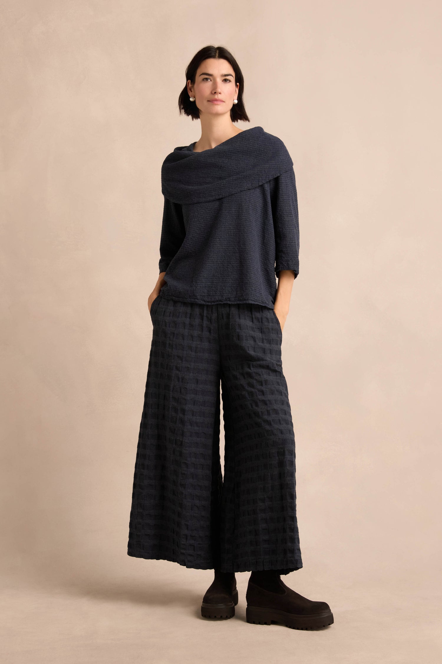 Woman stands against a beige background wearing a Navy Draped Neck Top in cotton-viscose jersey, wide-leg textured pants, and black shoes, with hands in her pockets for a relaxed fit look.
