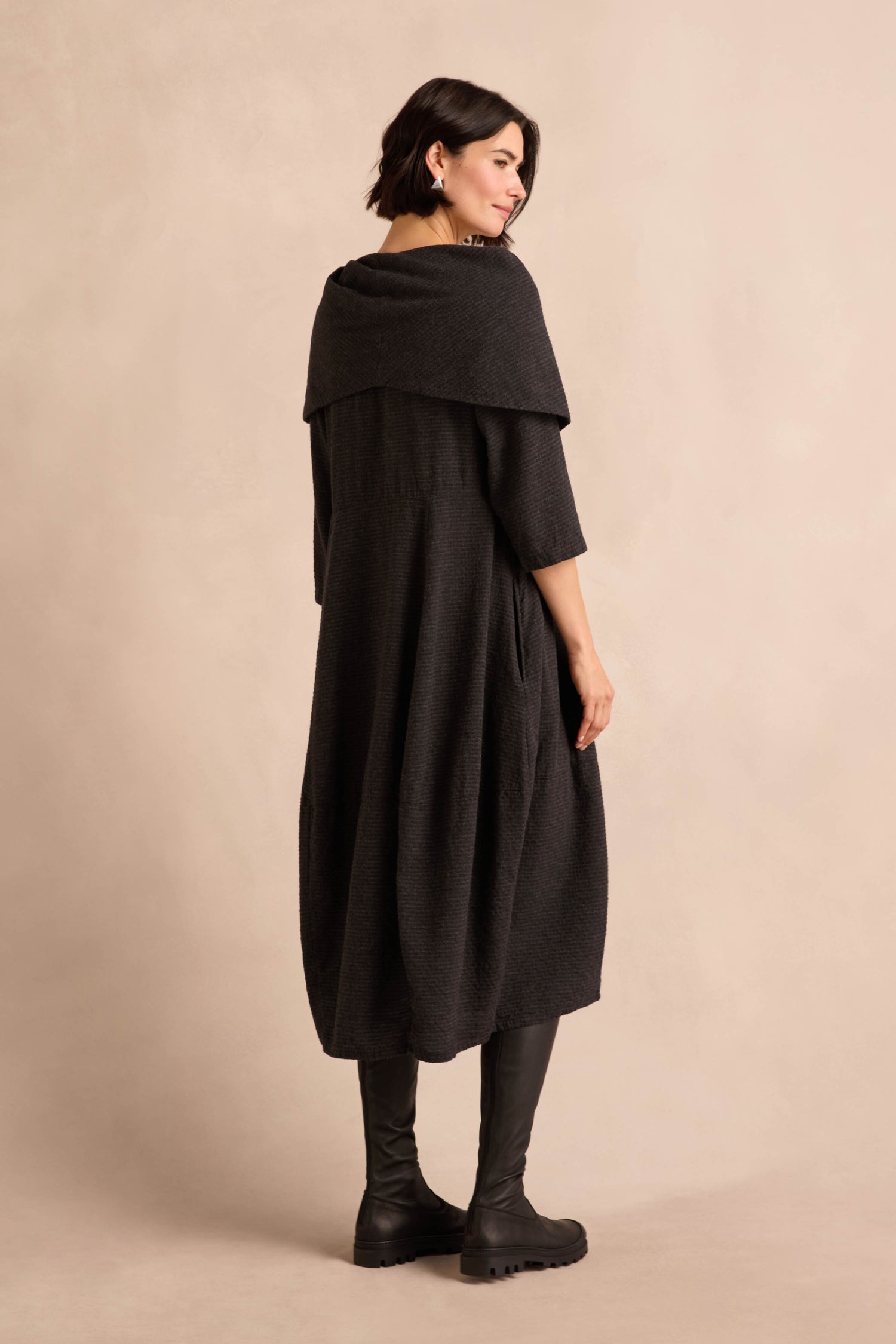 A person stands with their back to the camera, wearing the Anthracite Draped Neck Bubble Dress— a dark, long, loose-fitting dress featuring a wide draped collar—paired with black boots against a beige background.