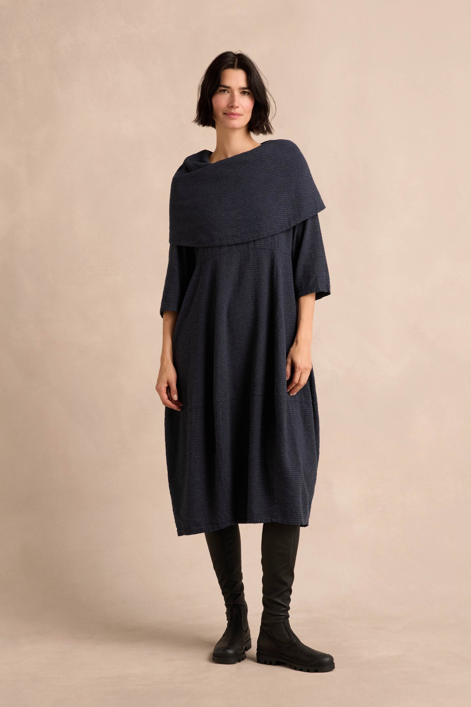 A person stands against a beige background wearing the Navy Draped Neck Bubble Dress, a loose-fitting piece crafted from soft cotton-viscose jersey, paired with black knee-high boots.