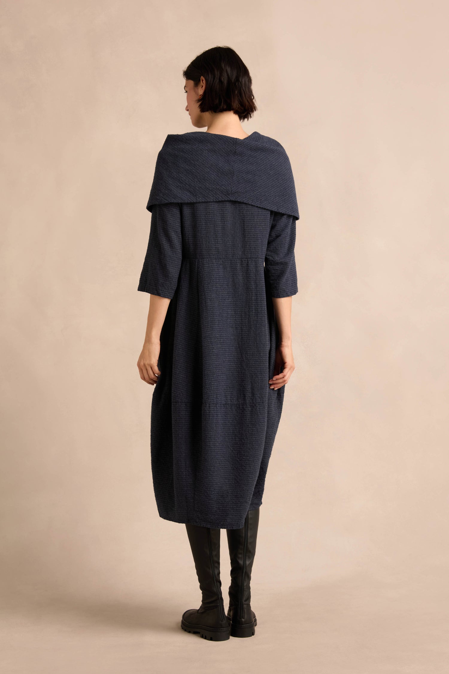 A person stands facing away, wearing the Navy Draped Neck Bubble Dress with a wide collar and knee-high black boots, against a plain beige background.
