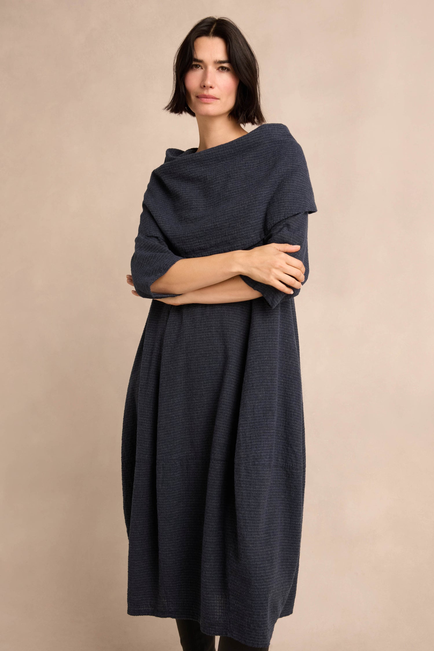 A woman with short dark hair wears the Navy Draped Neck Bubble Dress—crafted from a dark blue cotton-viscose jersey with a subtle bubble silhouette—paired with a matching shawl, standing against a neutral beige background with her arms crossed.