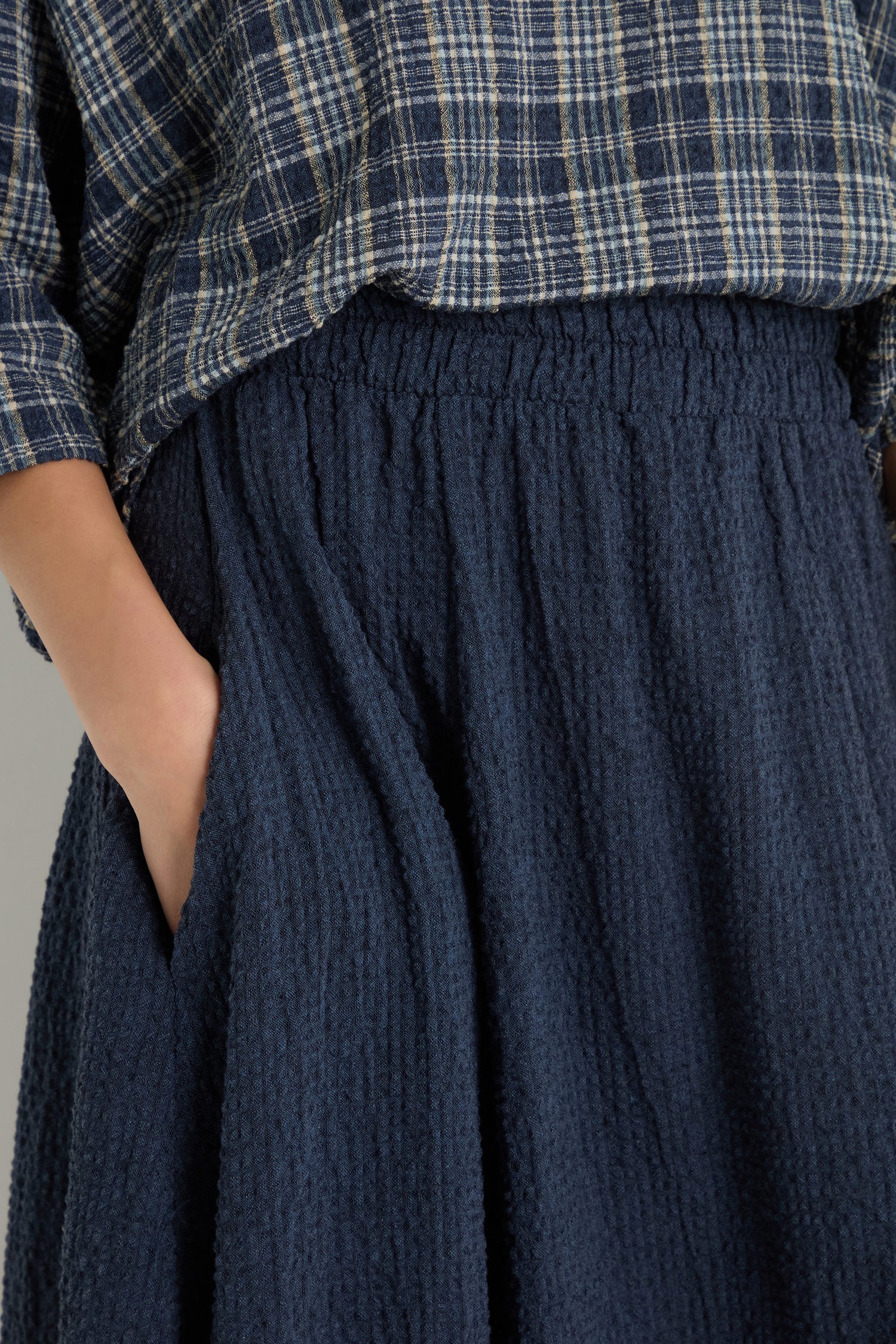 A person models the Crinkle Bubble Skirt, featuring an elasticated waist, paired with a blue plaid shirt and one hand in the pocket.