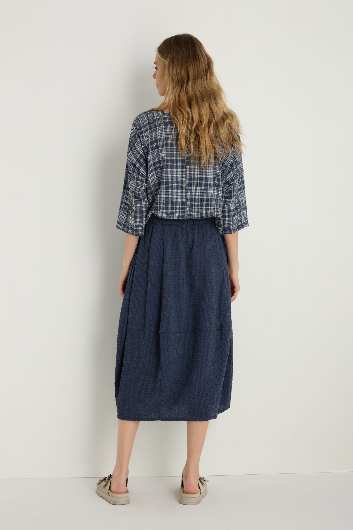 A person stands with their back to the camera, wearing a blue plaid shirt and the Crinkle Bubble Skirt in navy with an elasticated waist, paired with beige shoes against a plain white background.