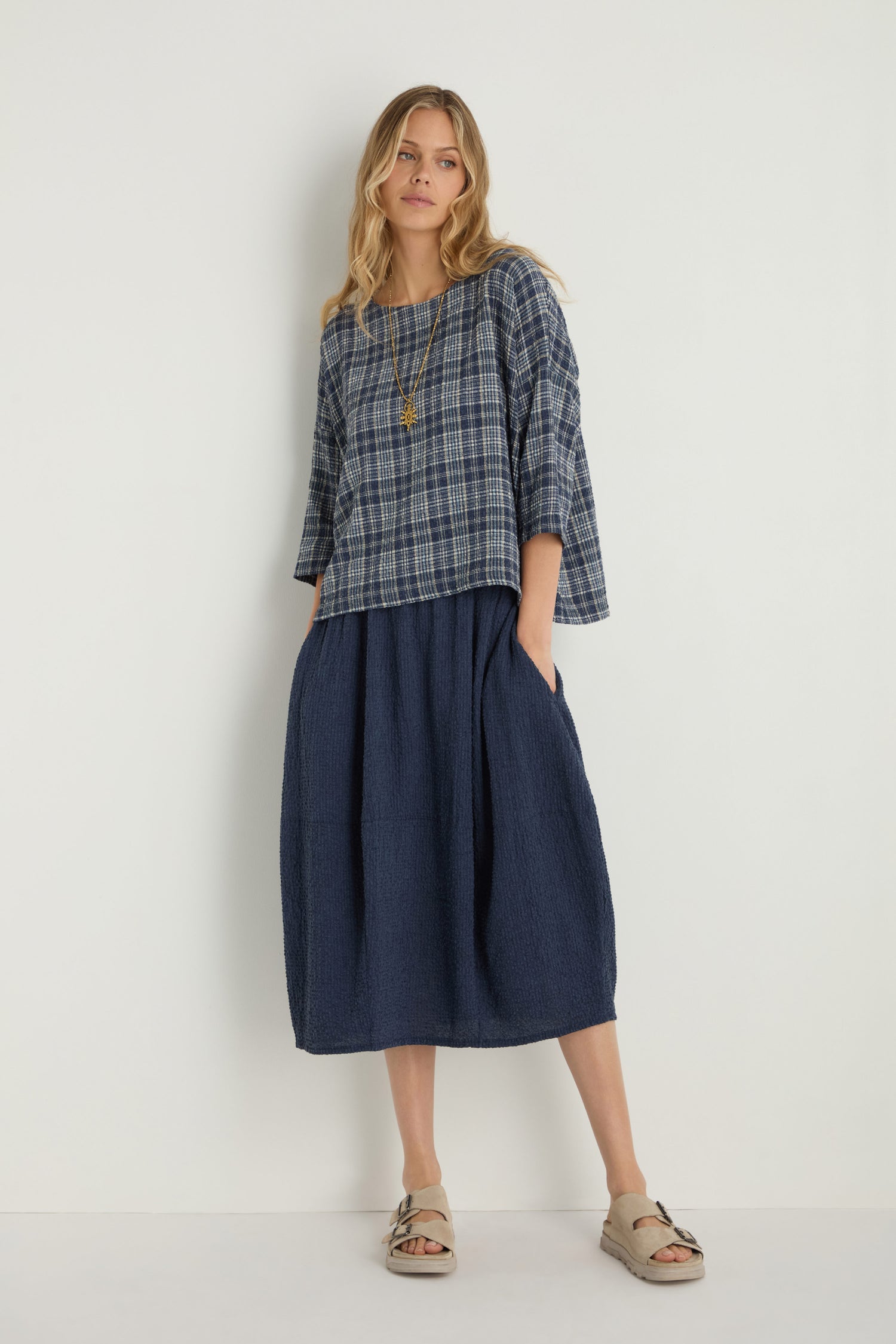 A woman stands against a plain background wearing the Check Boxy Top, a blue plaid boxy top in textured cotton blend, paired with a navy midi skirt, beige sandals, and a long necklace.