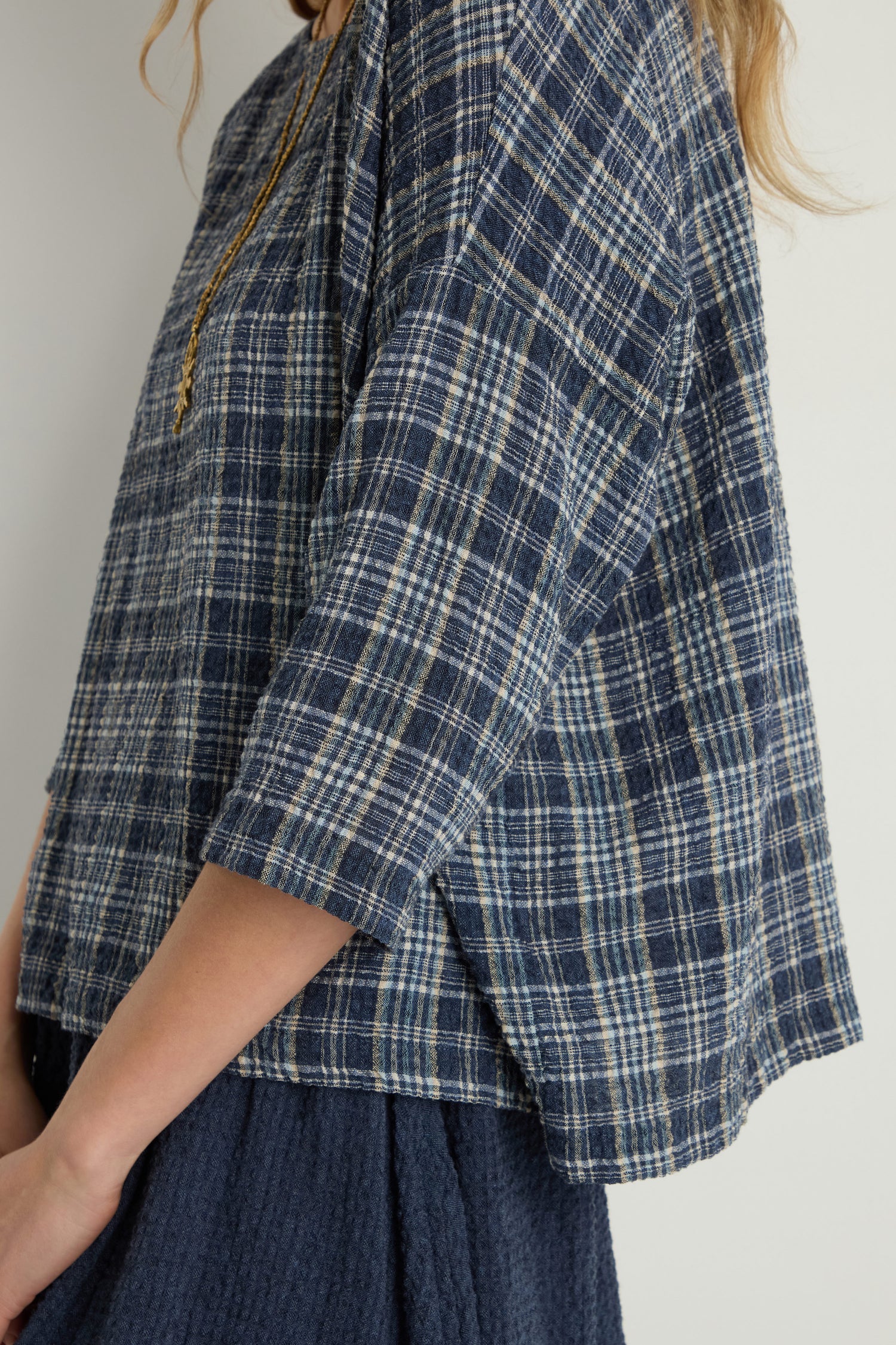 A person wears the Check Boxy Top in blue and white plaid with side slits, paired with a textured navy skirt. Only the upper body is visible.