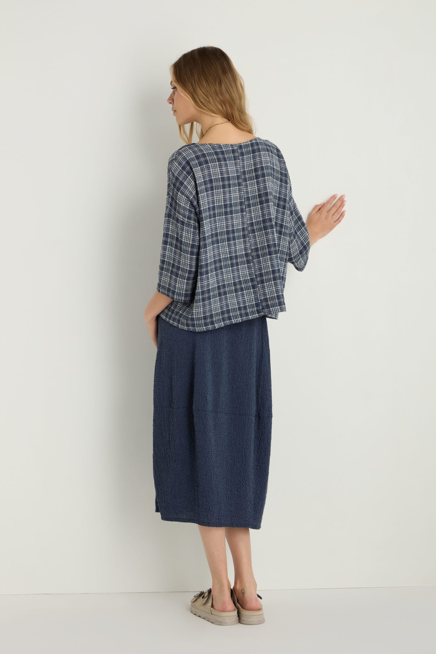 A woman stands against a white wall, facing away, wearing the Check Boxy Top in blue plaid, a navy midi skirt, and beige slip-on shoes.