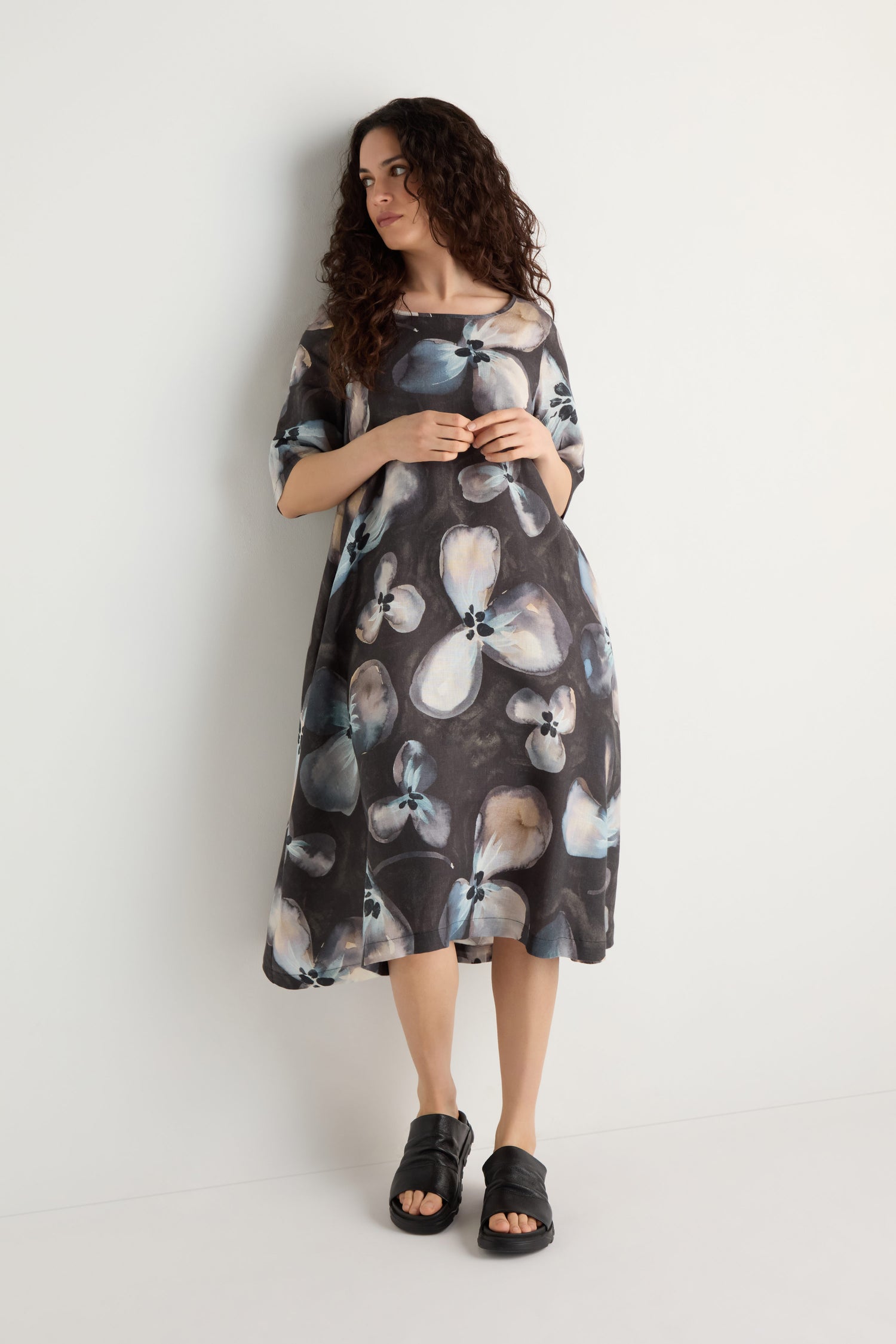 Woman with long curly hair wearing the Abstract Floral Linen Flared Dress and black sandals, standing against a plain white wall.